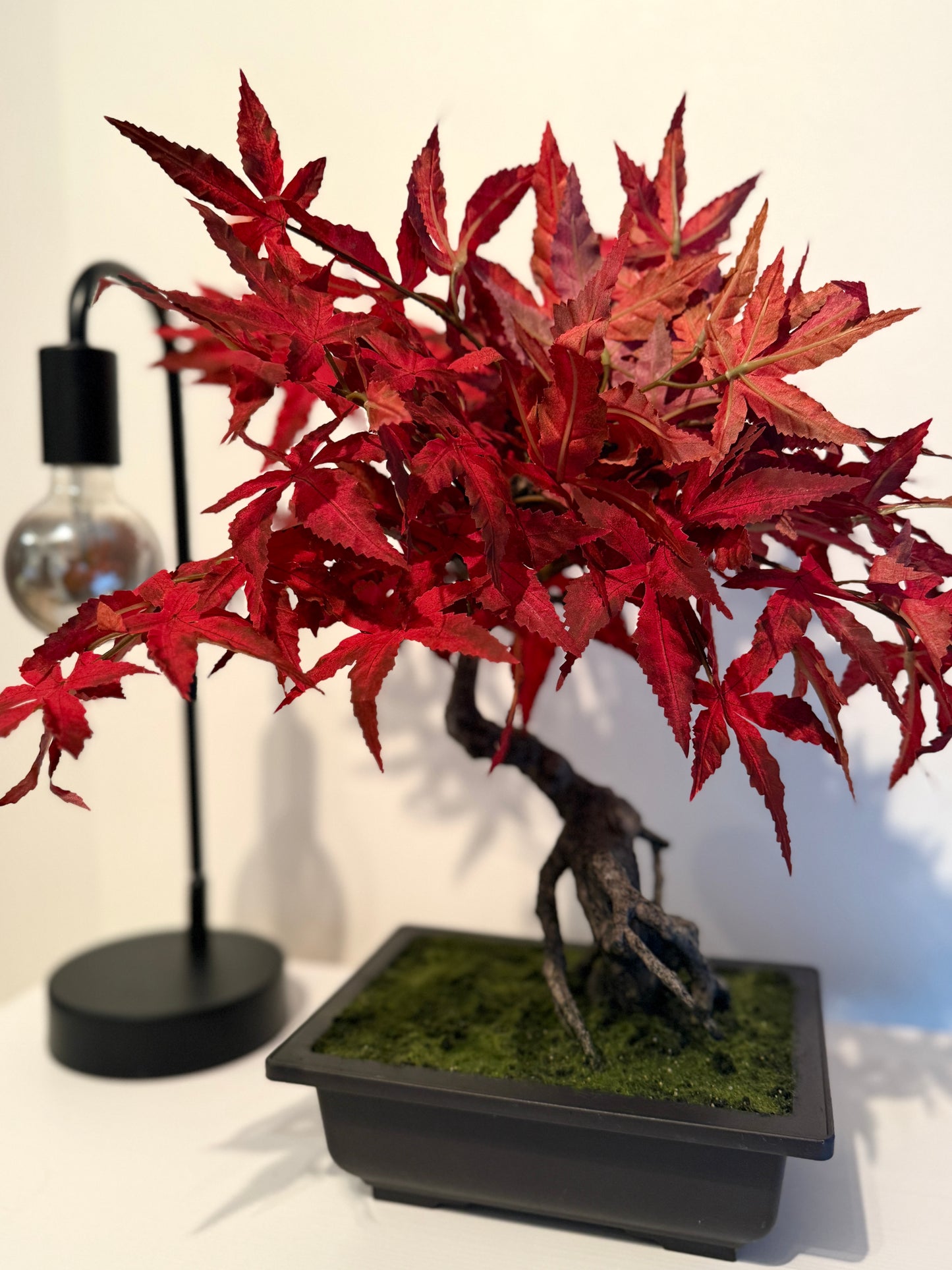 Front view of a Plantish premium artificial red maple bonsai, featuring vibrant red leaves and a realistic textured stem, placed on top of a white dresser next to a minimalist black lamp.