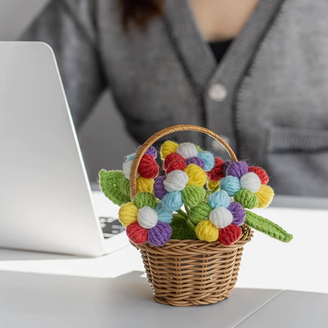 A close up image of the Plantish crocheted flower wicker basket with multicoloured flowers, placed on a white table with a white wall in the background.