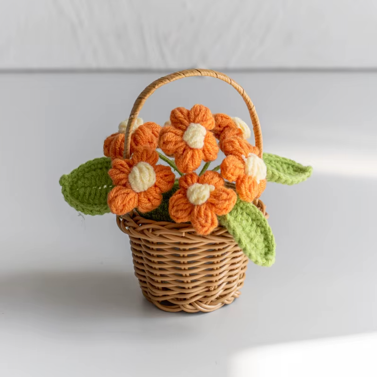 A close up image of the Plantish crocheted flower wicker basket with orange flowers, placed on a white table with a white wall in the background.