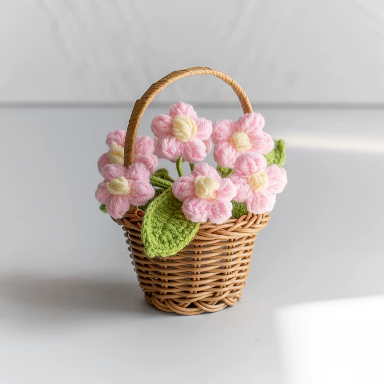 A close up image of the Plantish crocheted flower wicker basket with pink flowers, placed on a white table with a white wall in the background.