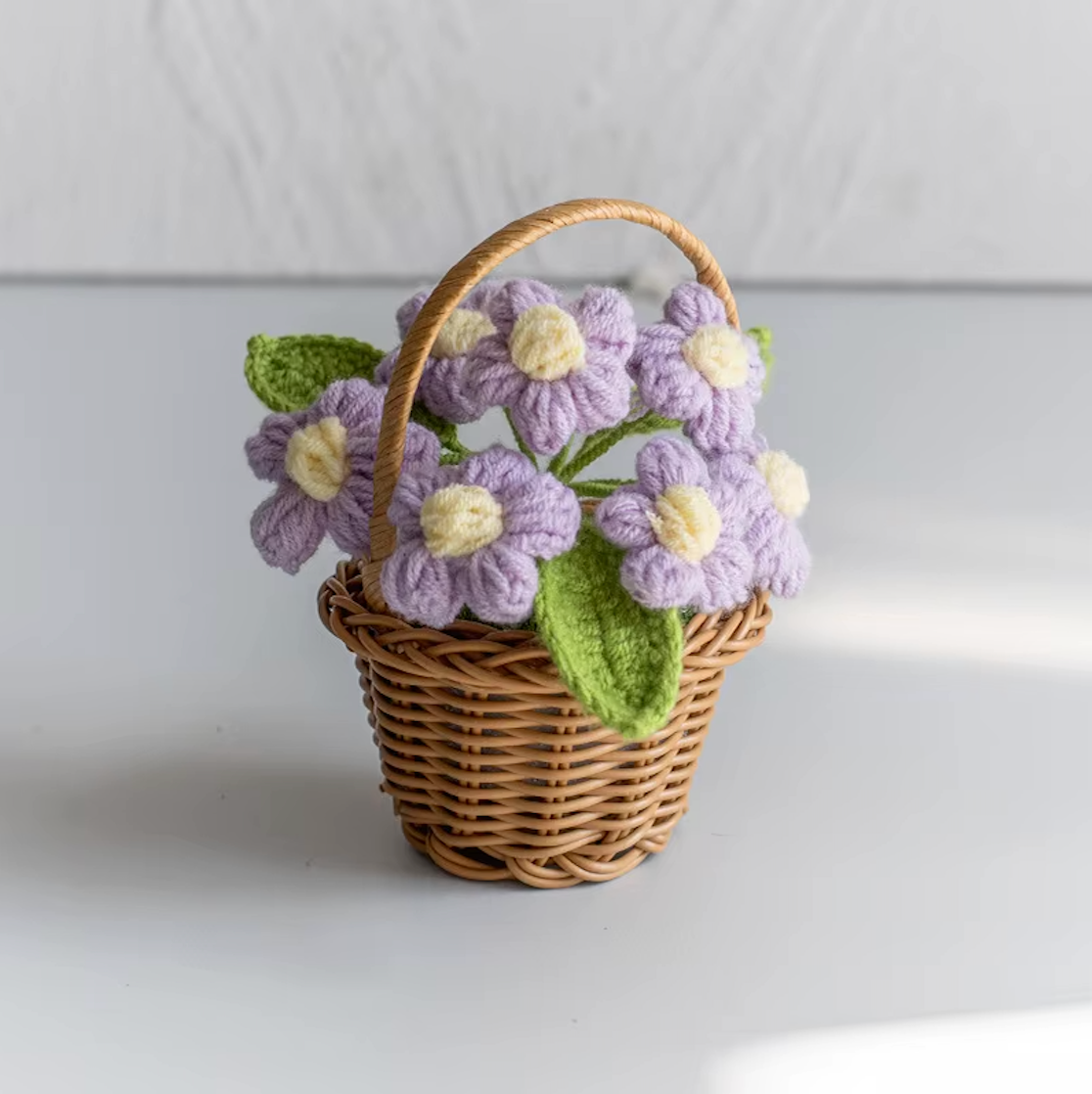 A close up image of the Plantish crocheted flower wicker basket with purple flowers, placed on a white table with a white wall in the background.