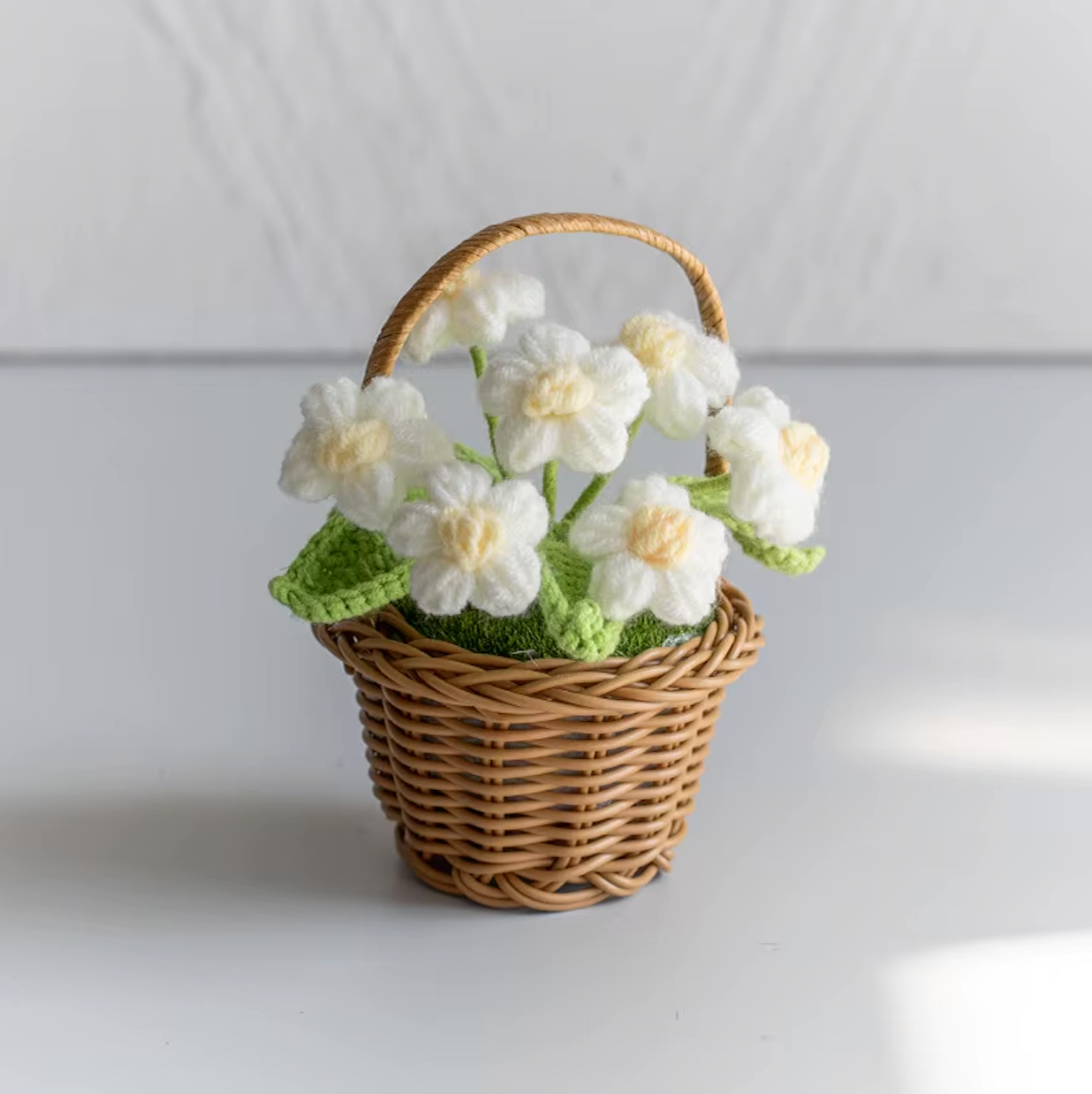 A close up image of the Plantish crocheted flower wicker basket with White flowers, placed on a white table with a white wall in the background.