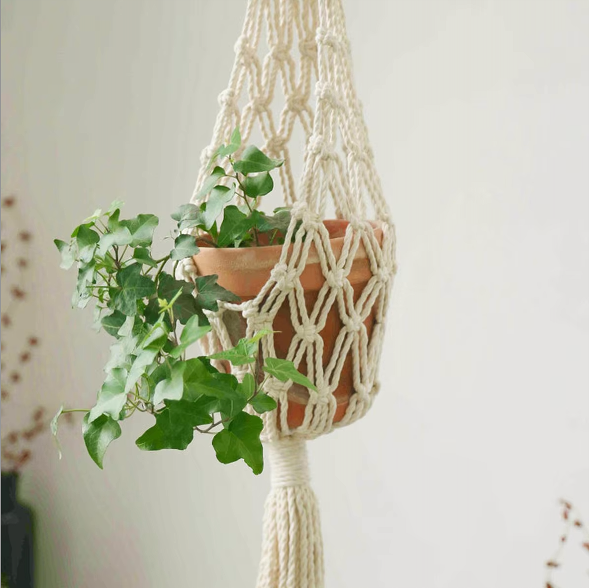 A close up of the Plantish Crosshatch Macrame, which has a terracotta plant pot with a green plant inside. The macrame is suspended in the corner of a bedroom.