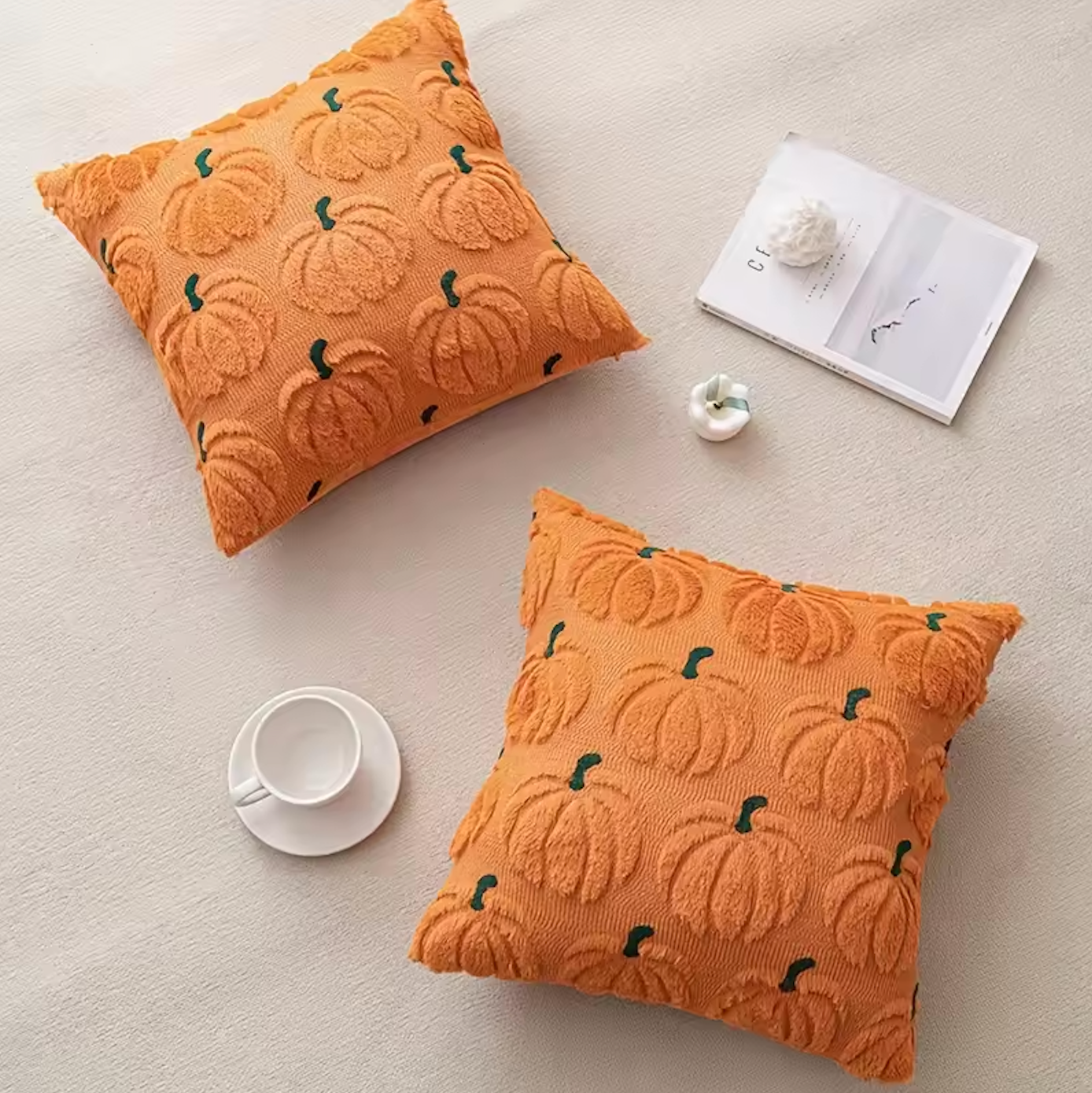 Two embroidered orange pillows with pumpkin patterns on a light surface with a cup and book.