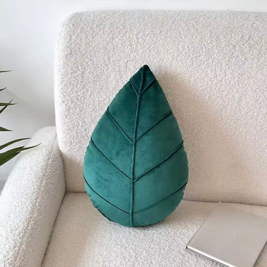 The Plantish Evergreen Plant Leaf Cushion in Dark Green postioned on a white sofa along with a book. The plush leaf detail is visible.