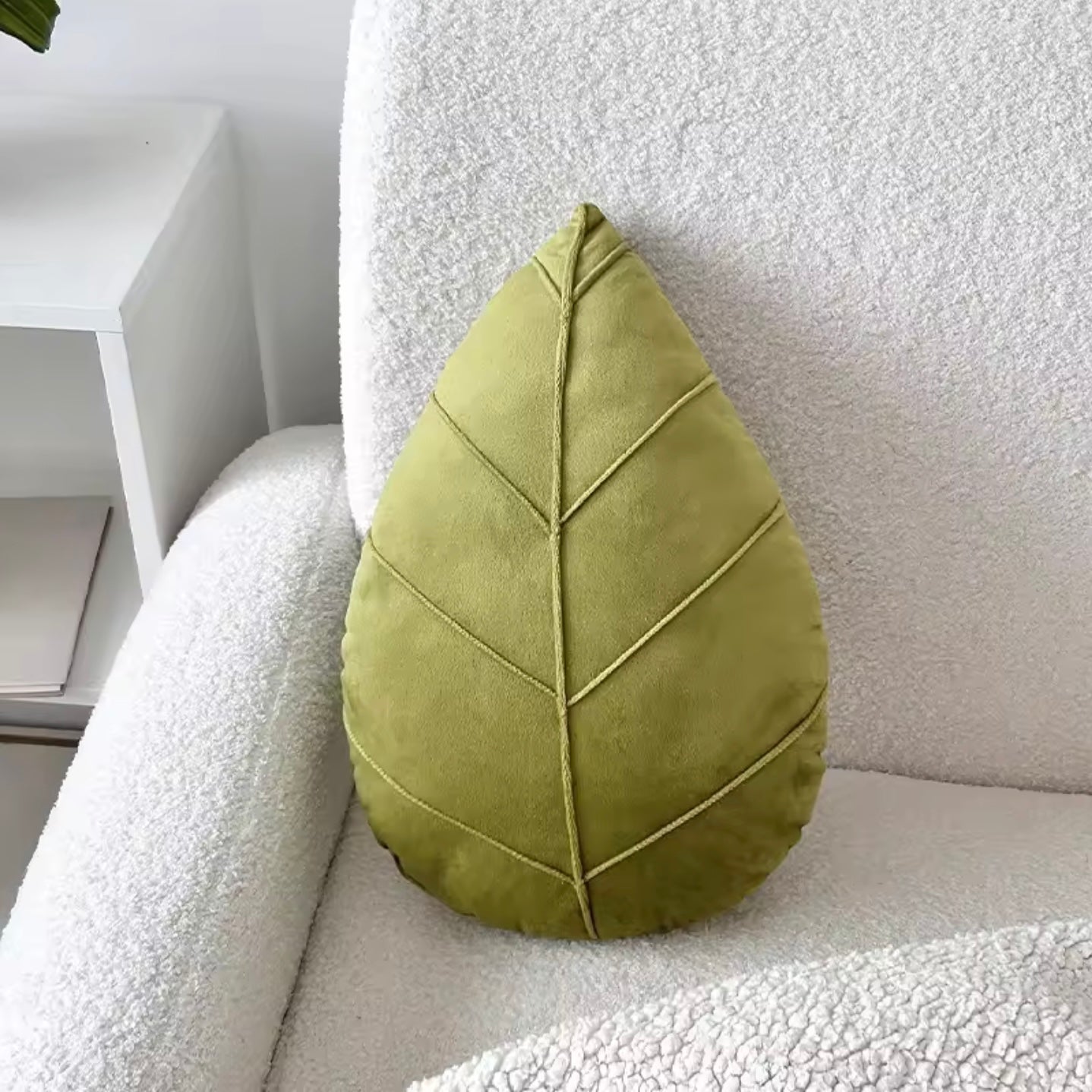 The Plantish Evergreen Plant Leaf Cushion in Light Green postioned on a white sofa. The plush leaf detail is visible, and there is a white bookcase next to the sofa