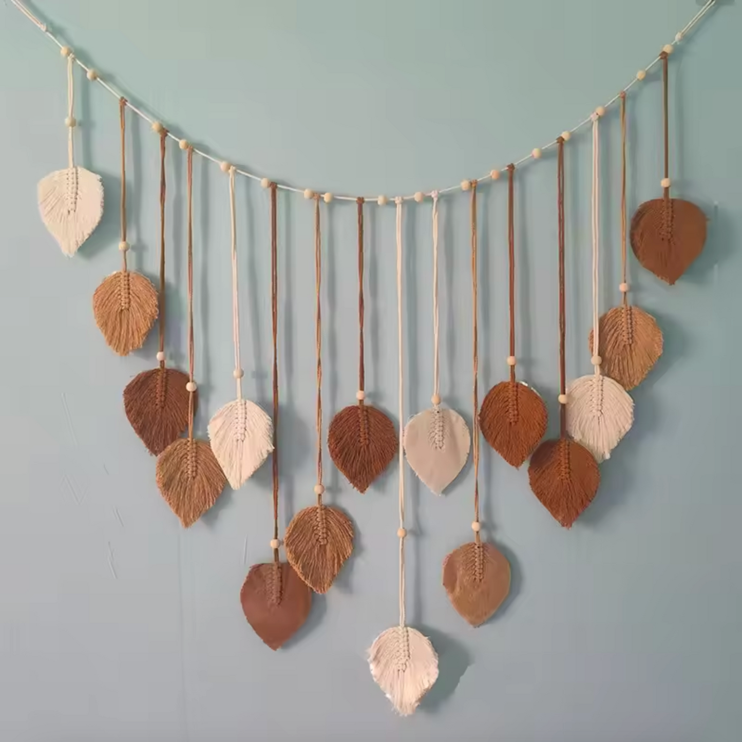 A Plantish Leaf Macrame with brown and white leaves and a white string hanging on a blue wall.