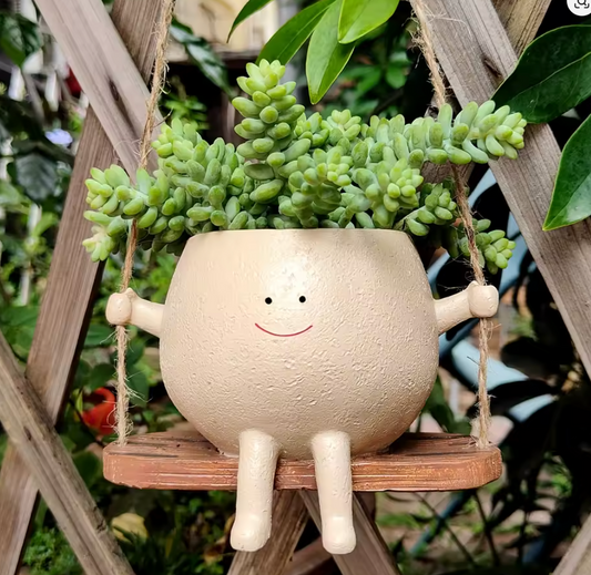 A close up of the Plantish Happy Swing Plant Pot, hanging in front of a wooden trellis in a garden. There is a green succulent inside the pot.