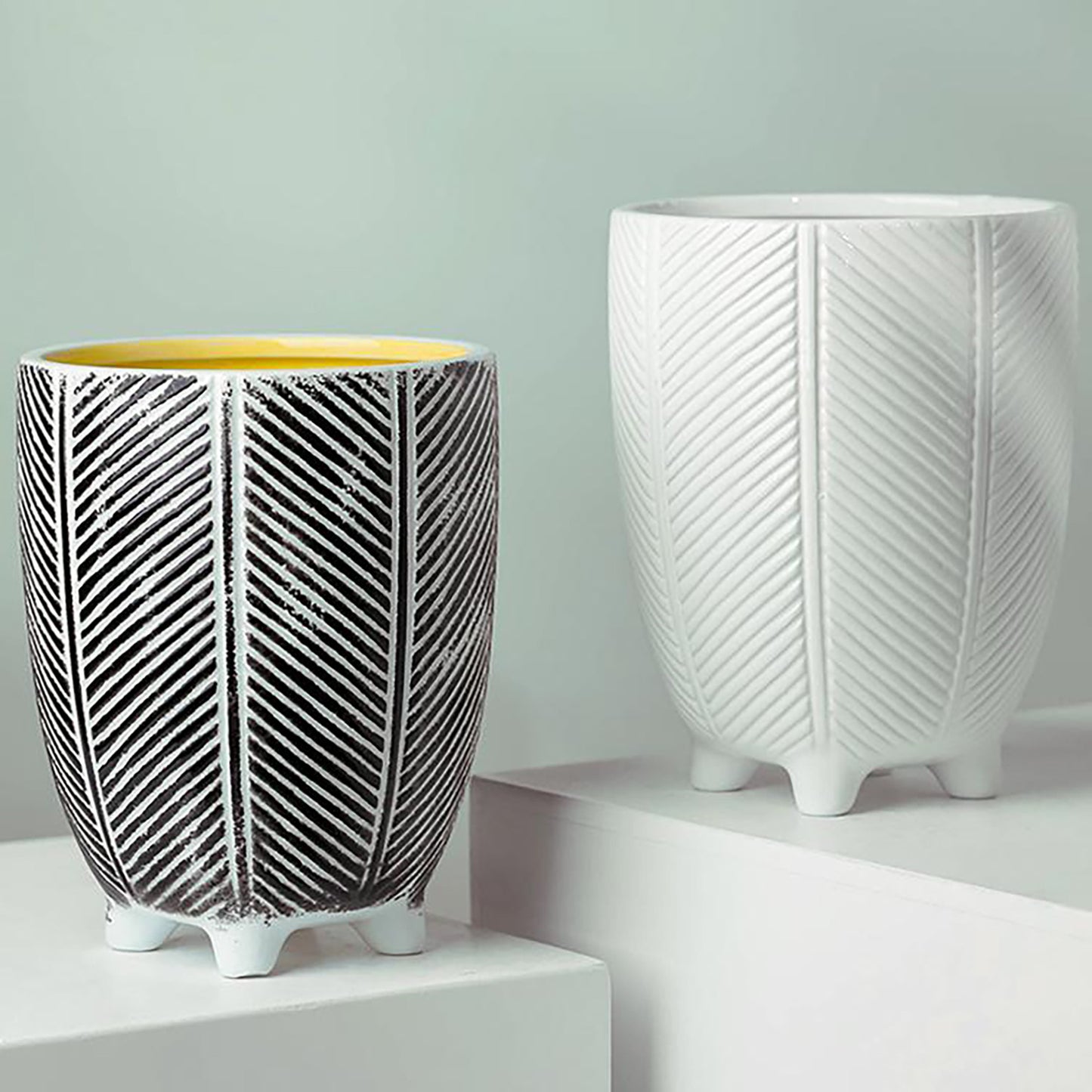 A view two ceramic plant pots with a zig zag herringbone design, the left one is black and white and the right one is white, both have four small feet.