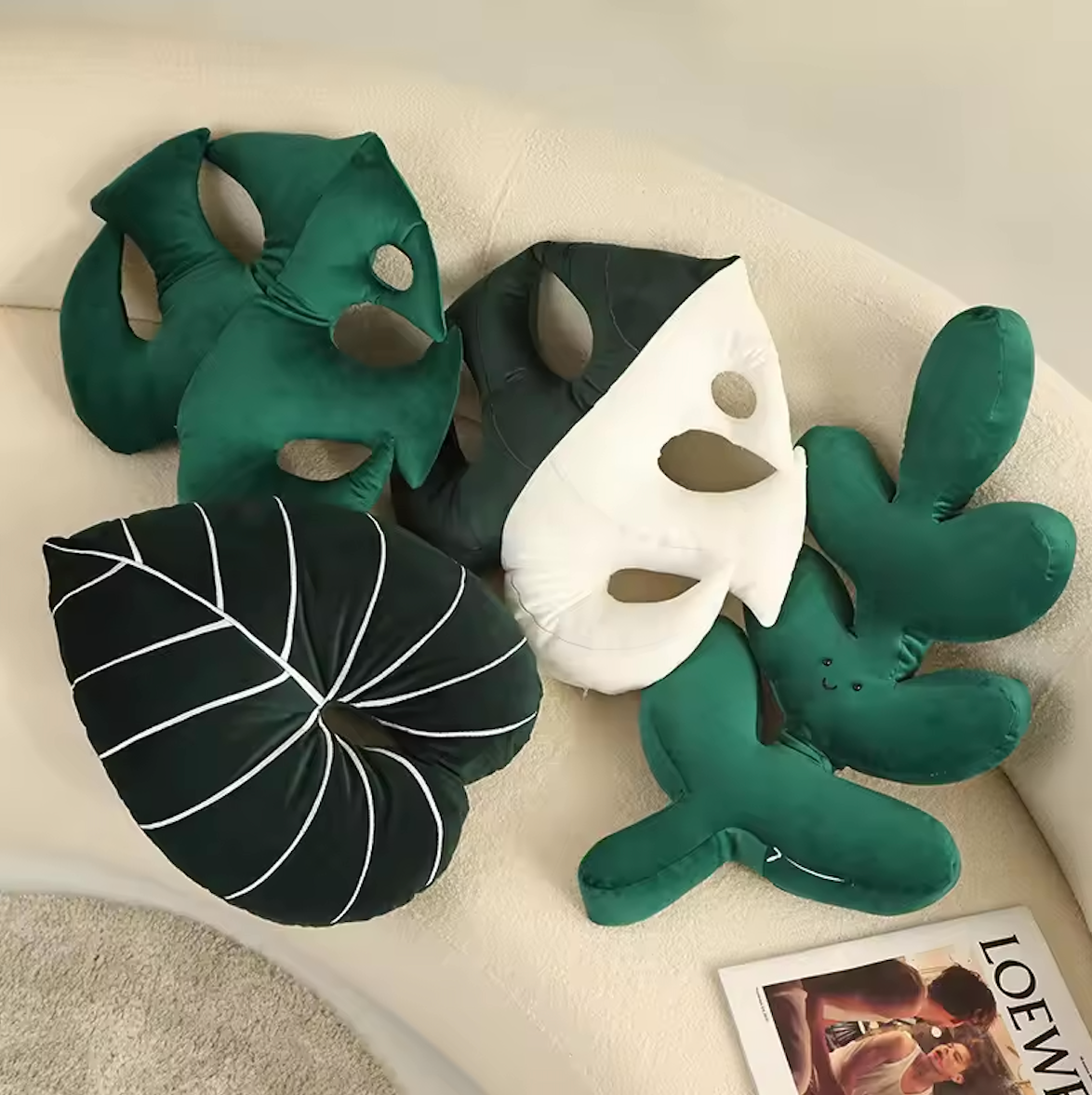 The Plantish Jungle Leaf Cushion in four different designs - two monstera leaf, one fern, one philodendron. Shown on a white sofa.