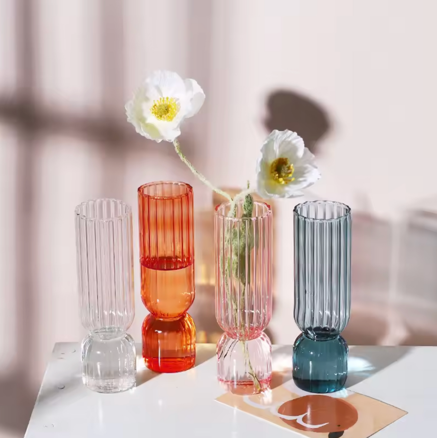 An image of the Plantish Little Nordic Vase in four colours - clear, light blue, orange, and pink. They are on a white table. There is a white flower inside the pink vase.