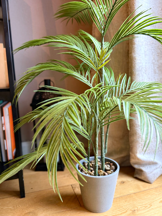 Back view of a Plantish artificial medium-sized palm, highlighting its lush green fronds and grey ceramic-look plastic pot with fixed pebbles, in the corner of a room next to a bookcase, with sunlight hitting the plant.