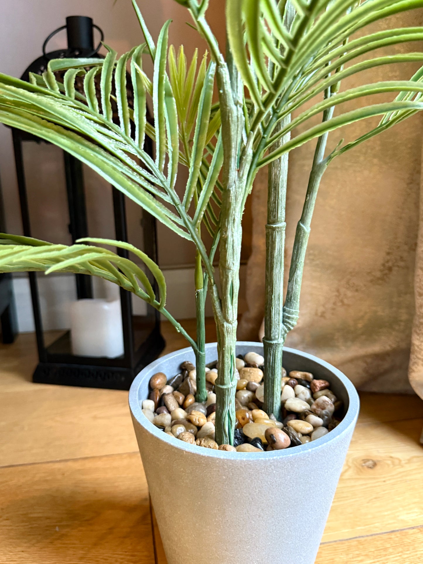 Close up of the Plantish artificial medium-sized palm, focusing on the realistic deep green fronds and the fixed pebbles in the grey ceramic-look plastic pot, with a black metal lantern encasing a white pillar candle in the background.