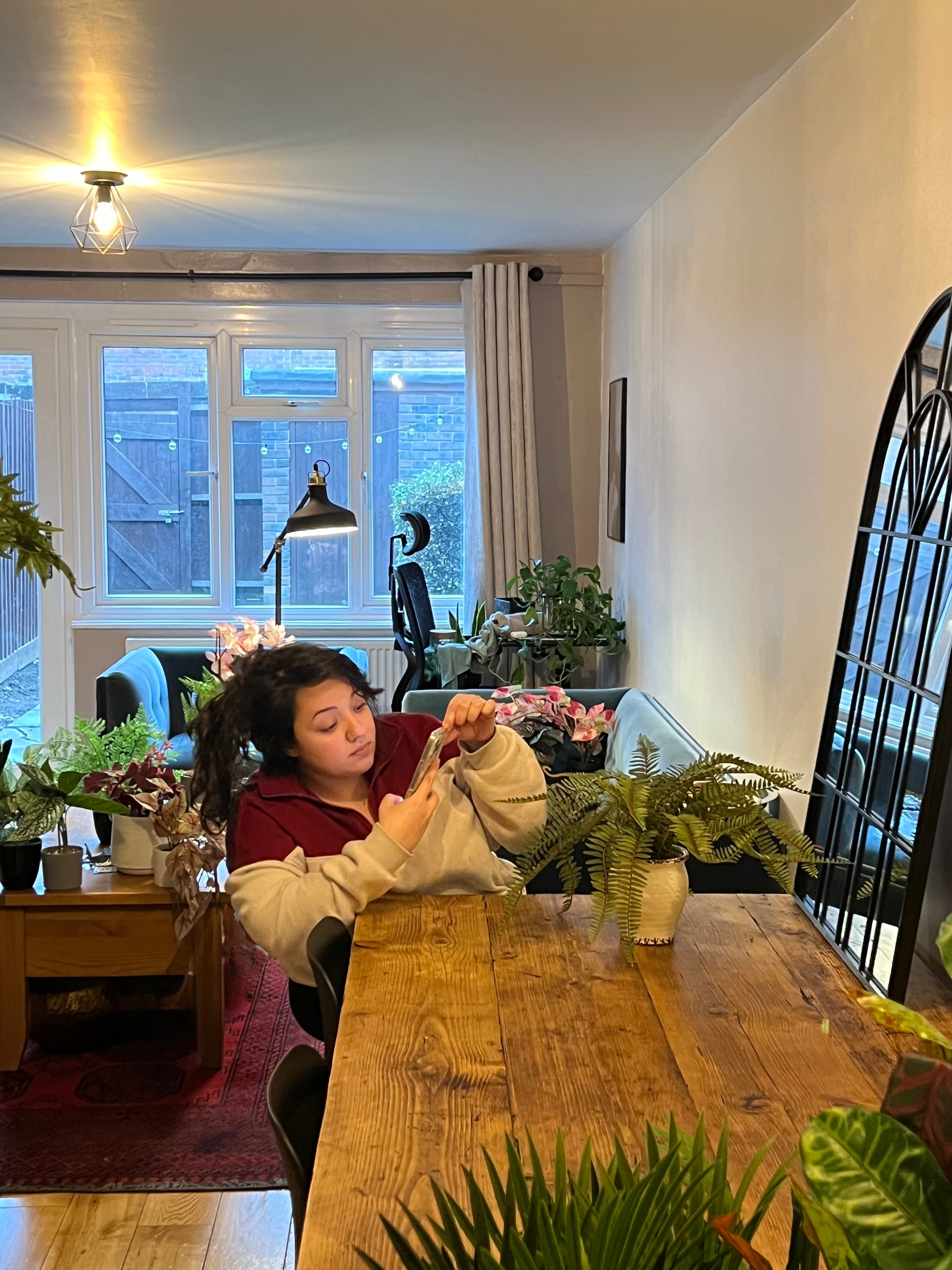 Person sitting at a wooden table with plants in a room with large windows.