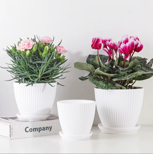 Three Plantish Ridged Plastic Planters in White, two with green flowers inside and on a white table, in front of a white wall.