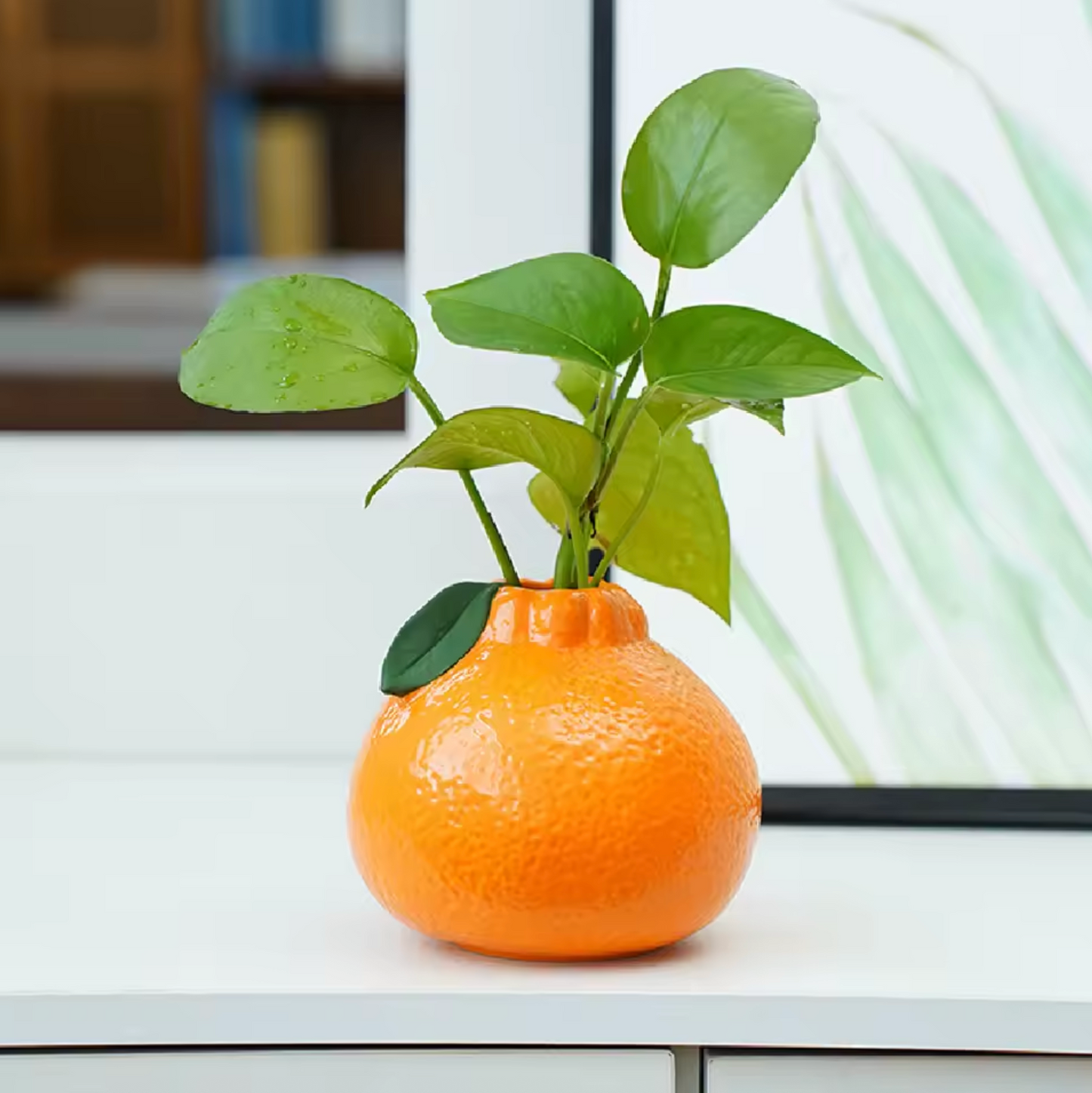 A profile view of the Plantish Tiny Orange Vase on a white surface. There is a green ivy plant stem inside the vase, and a black picture frame in the background.