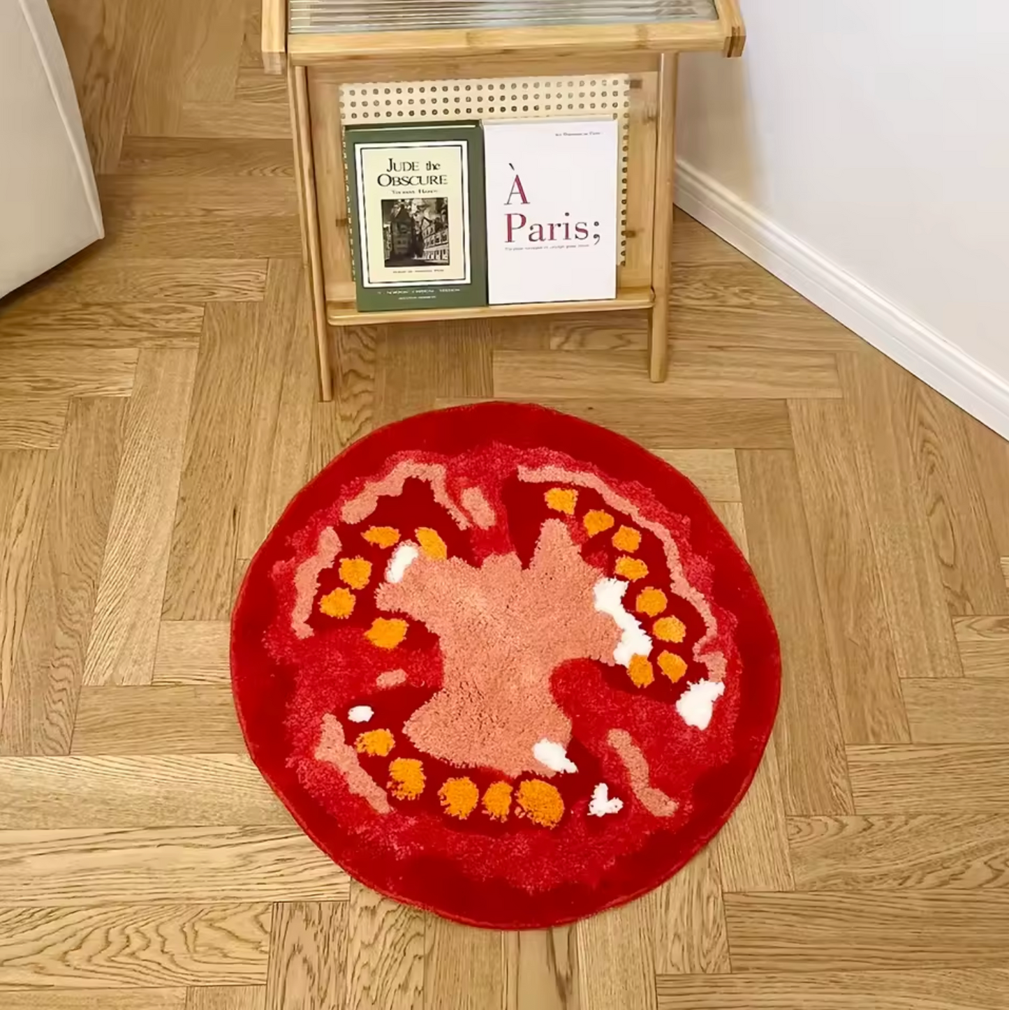 The Plantish Tomato Tufted Rug placed on a herringbone wooden floor, with a rattan magazine holder positioned behind that has two books on it.