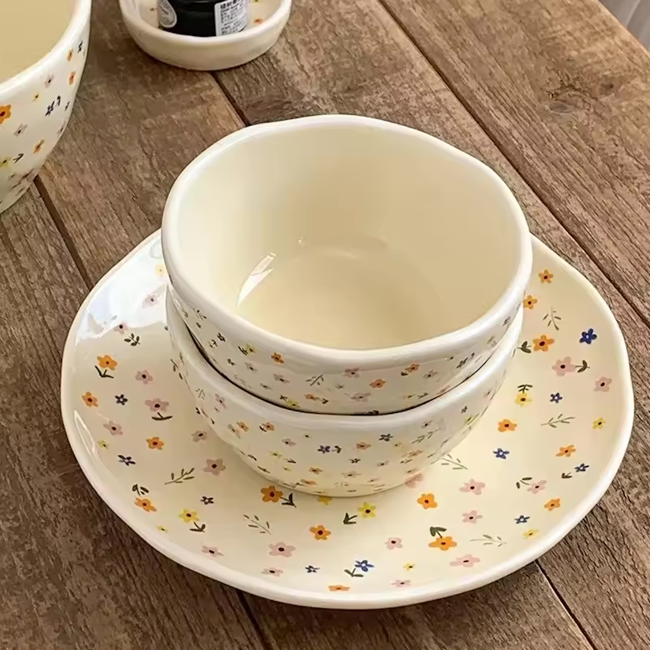 Two Plantish Wildflower small bowls stacked on one plate. Other items of the collection are visible in the background. They're all on a wooden table.