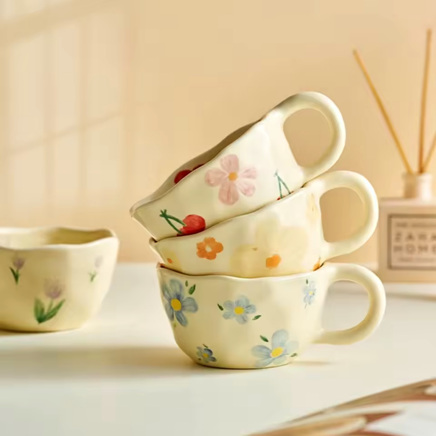 A close up of three Plantish Wonky Flower Mugs in Cherry Blossom, Blue Flower, and Orange Flower in a stack. The Purple Tulip variant is in the background and all are on a white table.