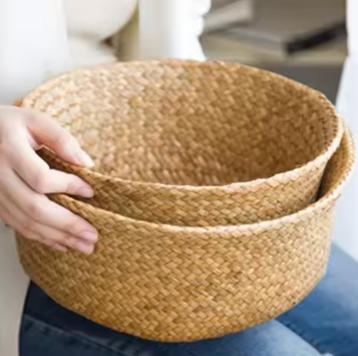 A close up of a lady holding two of the Plantish Woven Wicker Basket, one inside the other and both are folded down to half their full height.