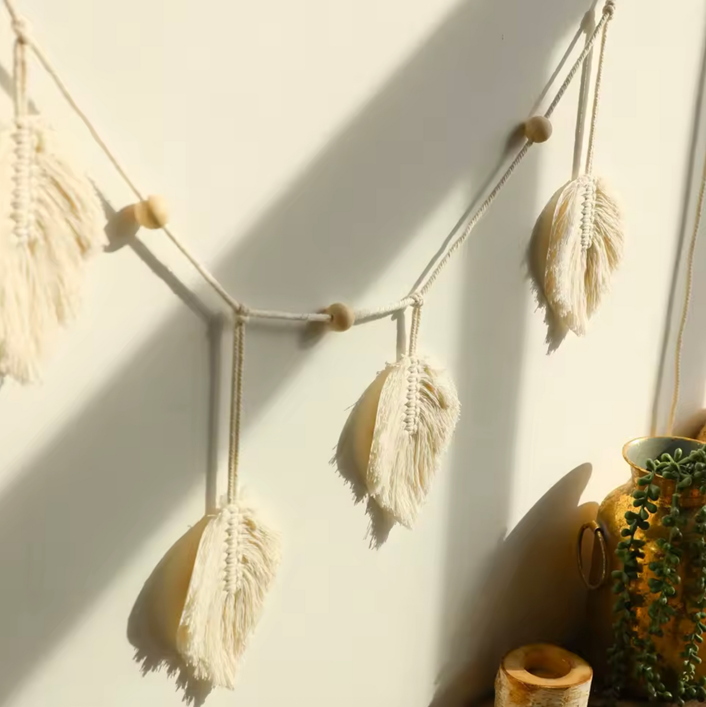 An angled view of the Plantish Bohemian White Leaf Macrame, hanging on a sunlit white wall with a green trailing plant visible to one side. 