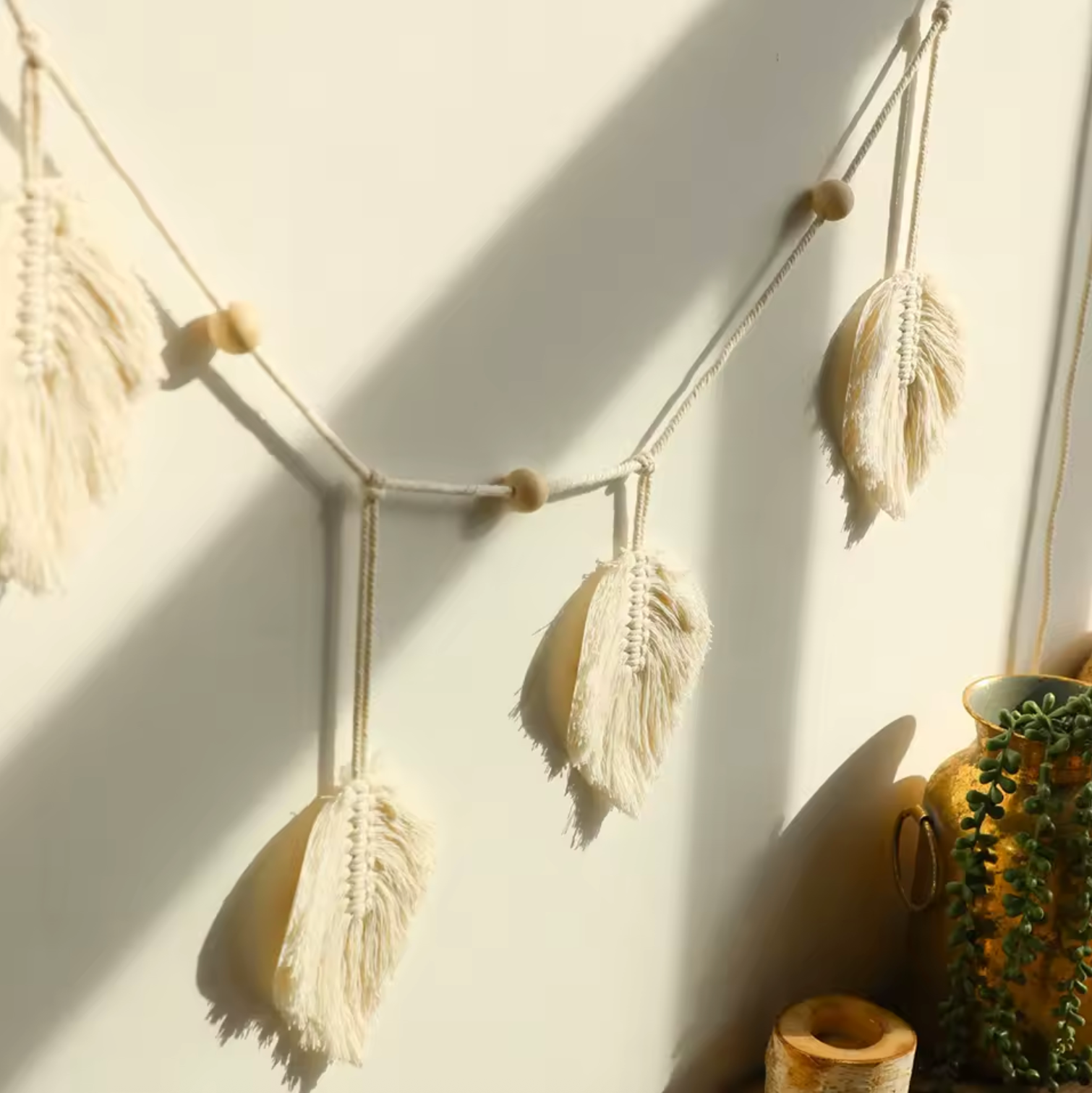 An angled view of the Plantish Bohemian White Leaf Macrame, hanging on a sunlit white wall with a green trailing plant visible to one side. 