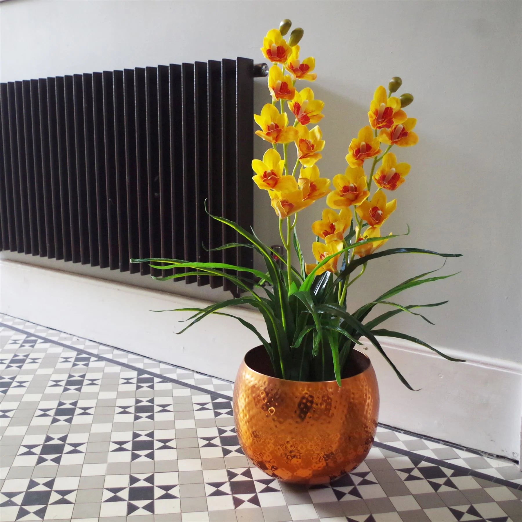 The Plantish artificial cymbidium orchid with yellow/orange flowers and green leaves, shown potted in a copper planter in a sunny hallway that has a mosaic floor. 