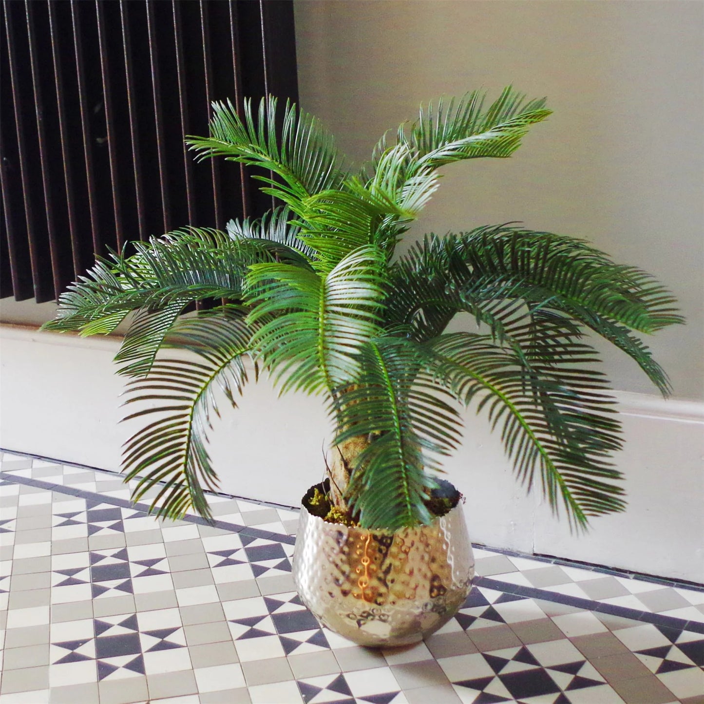 The Plantish artificial cycas palm shown in a decorative silver pot and placed in a sunlit hallway with a mosaic flooring. The green leaves and trunk are visible.