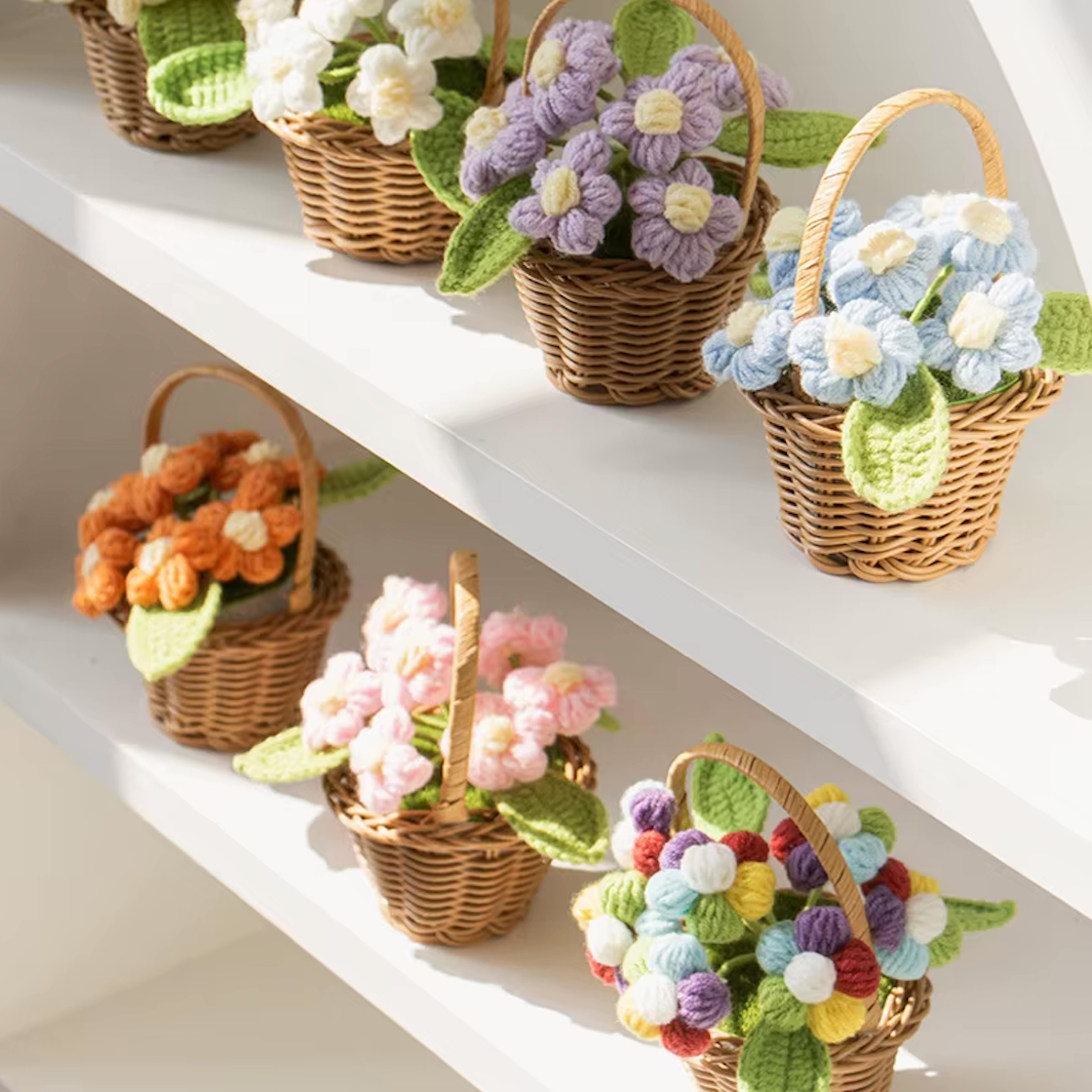 An image of all six Plantish crocheted flower wicker baskets placed on two white shelves, with three on each shelf. Sunlight is hitting the lower shelf.