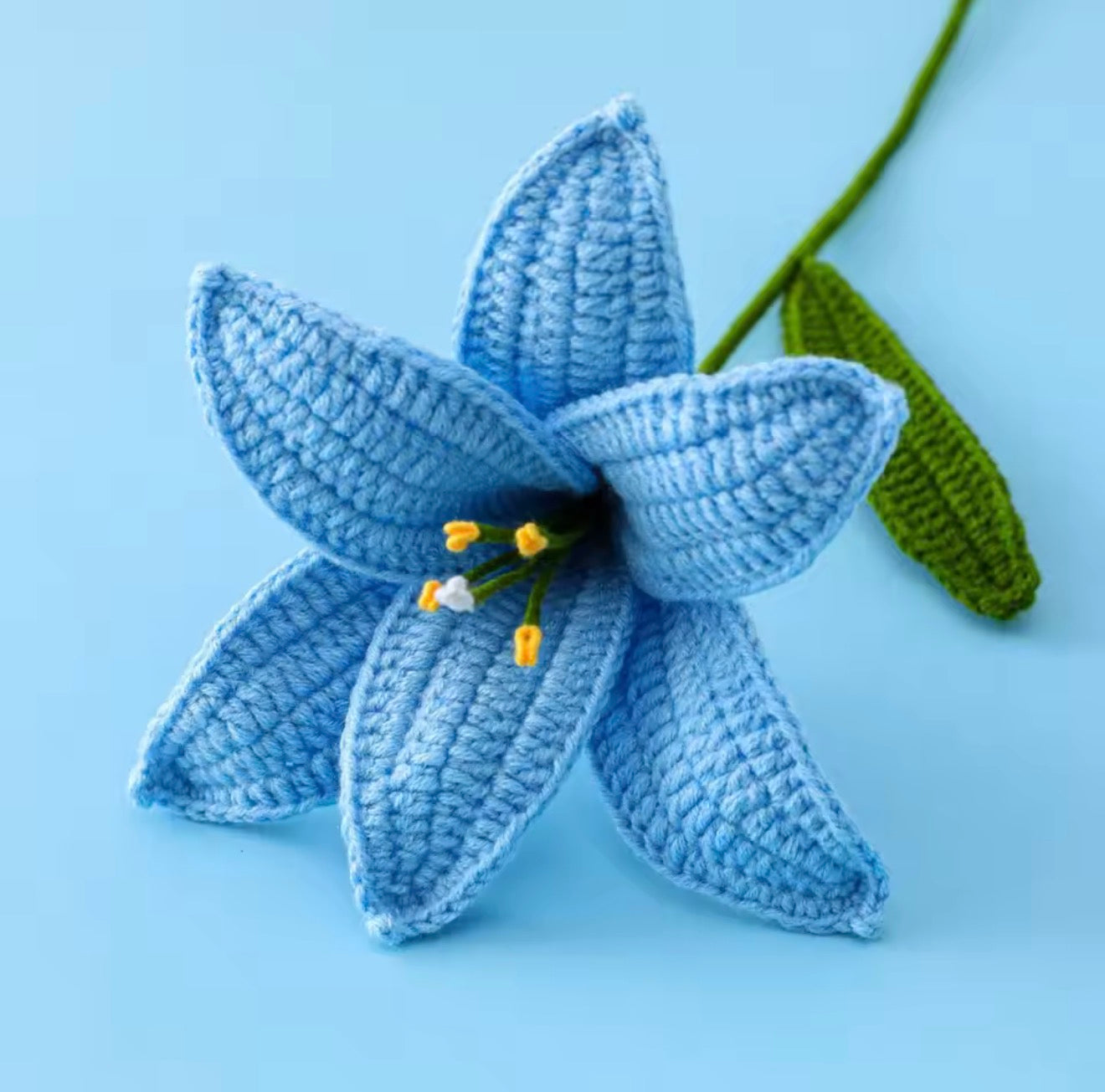 A close up of the top of the Plantish Crocheted Lily in Blue, showing off the detailed petals and green stem against a blue background.
