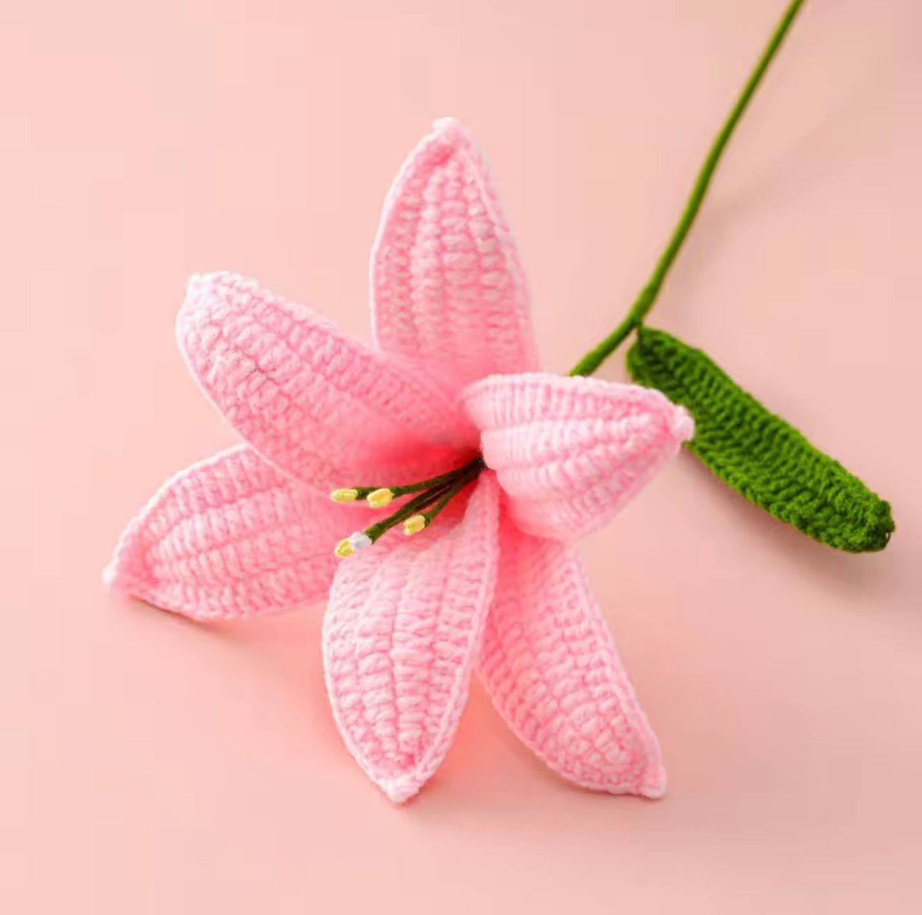 A close up of the top of the Plantish Crocheted Lily in Pink, showing off the detailed petals and green stem against a pink background.