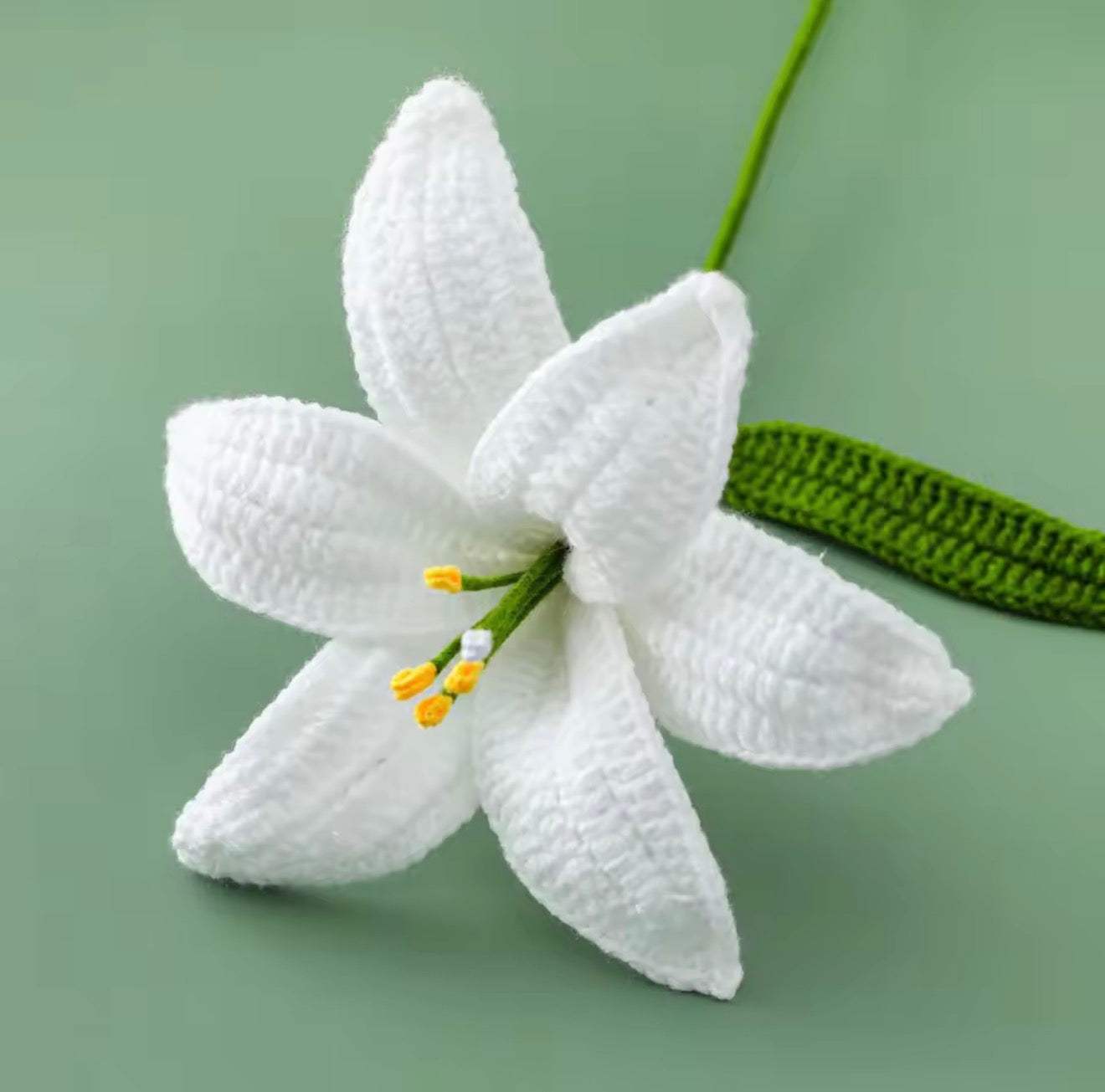 A close up of the top of the Plantish Crocheted Lily in White, showing off the detailed petals and green stem against a green background.

