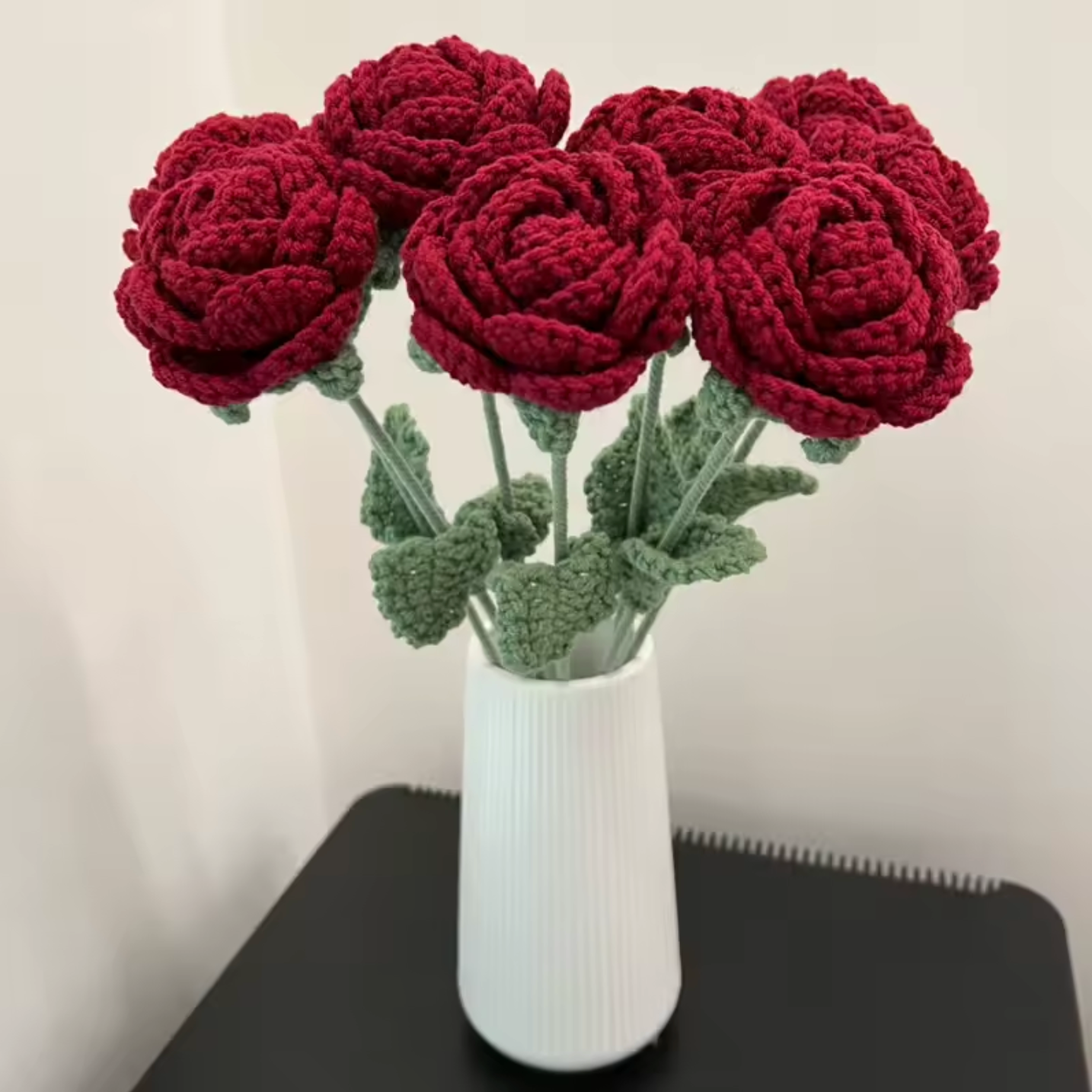 Six Plantish Crocheted Red Roses in a white ceramic vase which has a vertical groove design. The red petals, green leaves and green stems visible. 