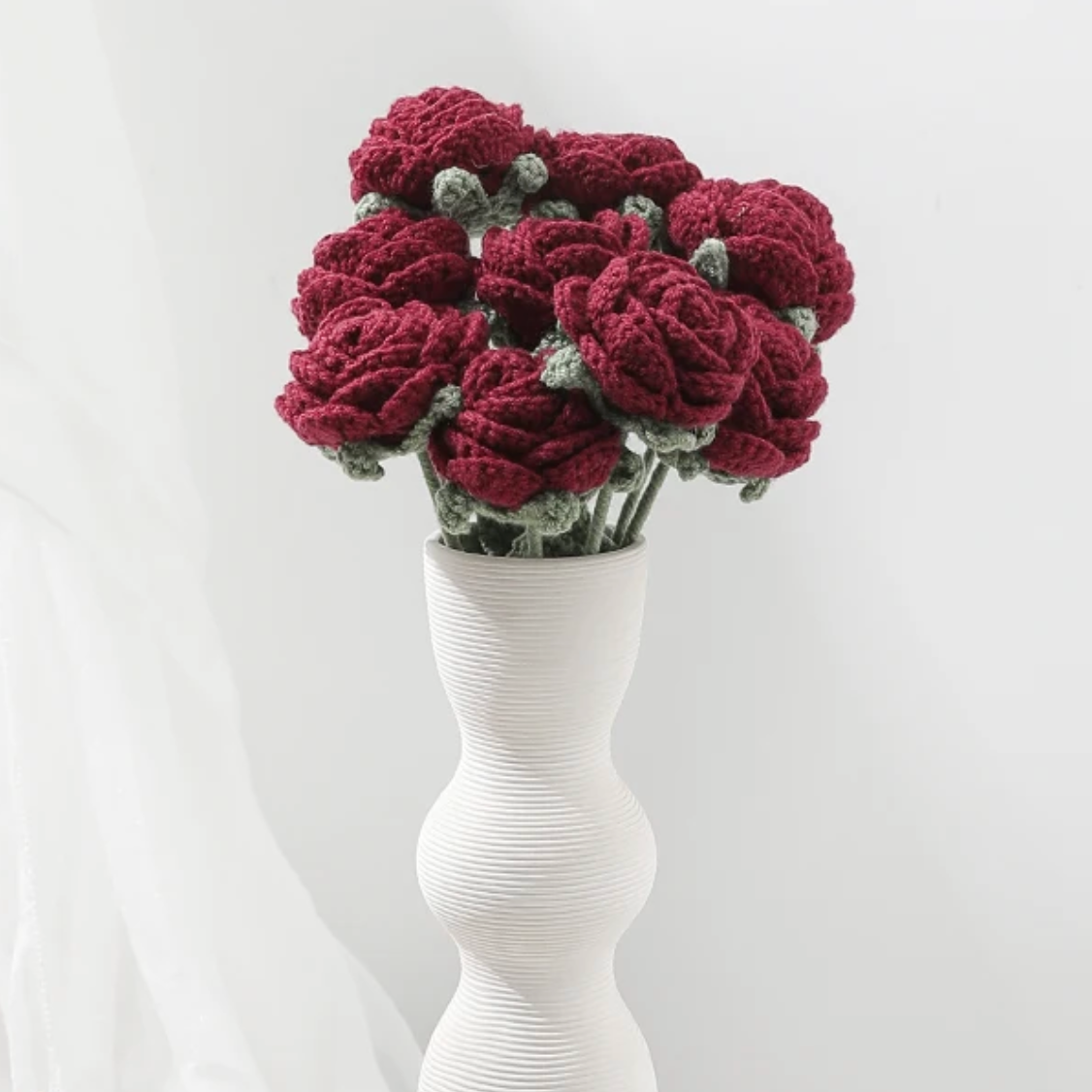 A bouquet of Plantish Crocheted Red Roses inside of a tall modern wavy white vase with horizontal groove design. It is shown against a white background.