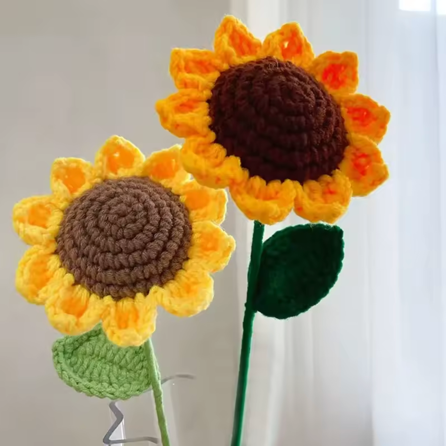 A close up of two Plantish Crocheted Sunflowers, showing the yellow and brown flower heads and green stems and leaves. 