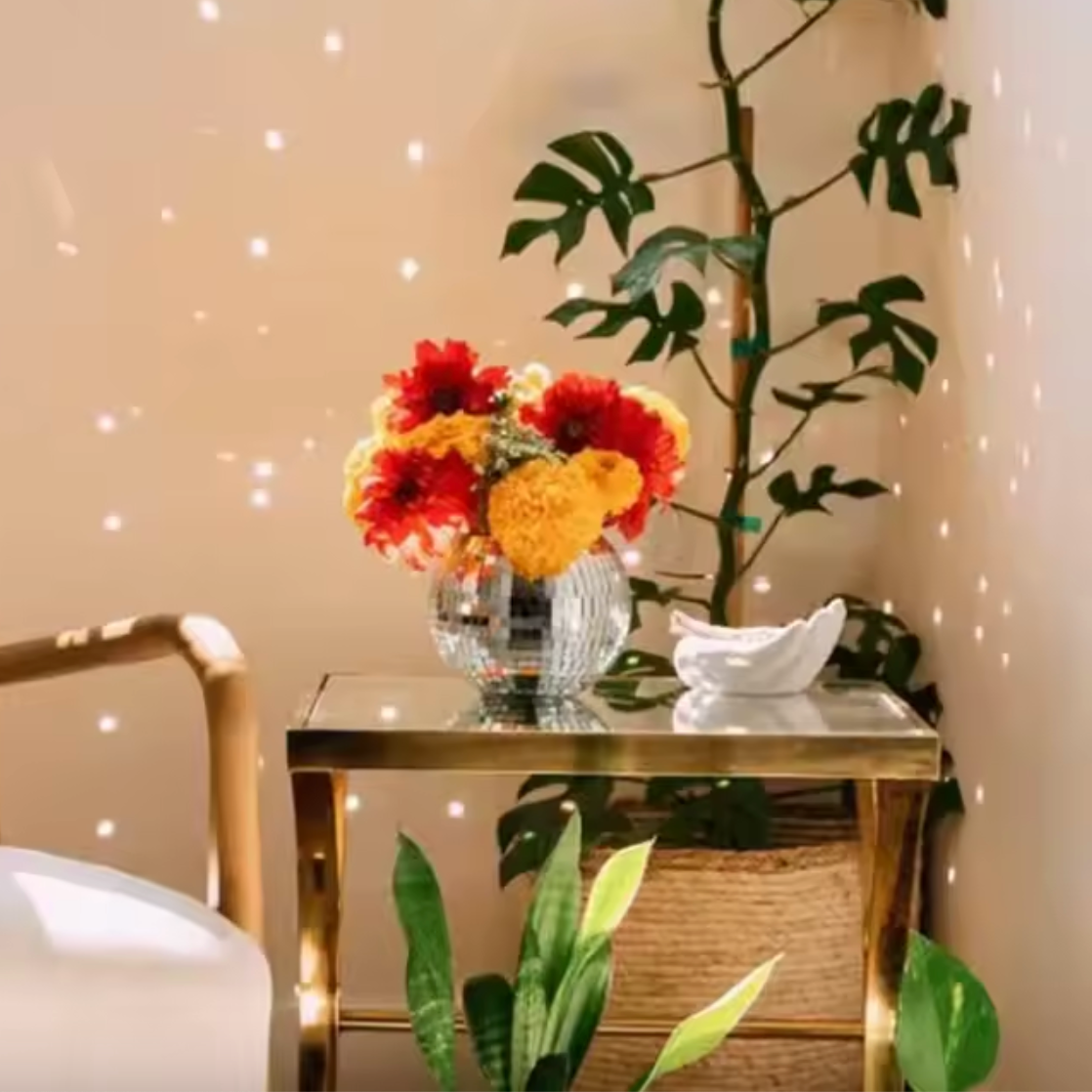 A close up image of the Plantish disco ball plant pot on a metal table, the sunlight reflecting off and hitting the walls. There are flowers in the disco ball pot. 