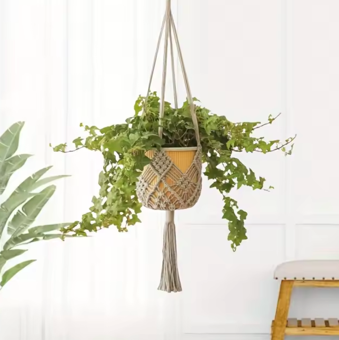 The Plantish Decorative Bohemian Macrame in Grey, hanging in a white living room holding a green plant in a yellow pot. There is another plant and a stool in the room. 
