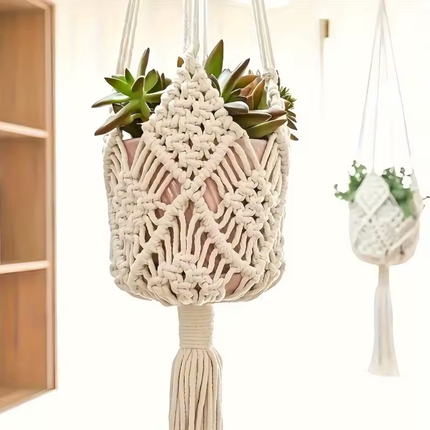 The Plantish Decorative Bohemian Macrame in White shown hanging in a sunlit room holding a pink pot with green succulents inside. There is another hanging in the background. 