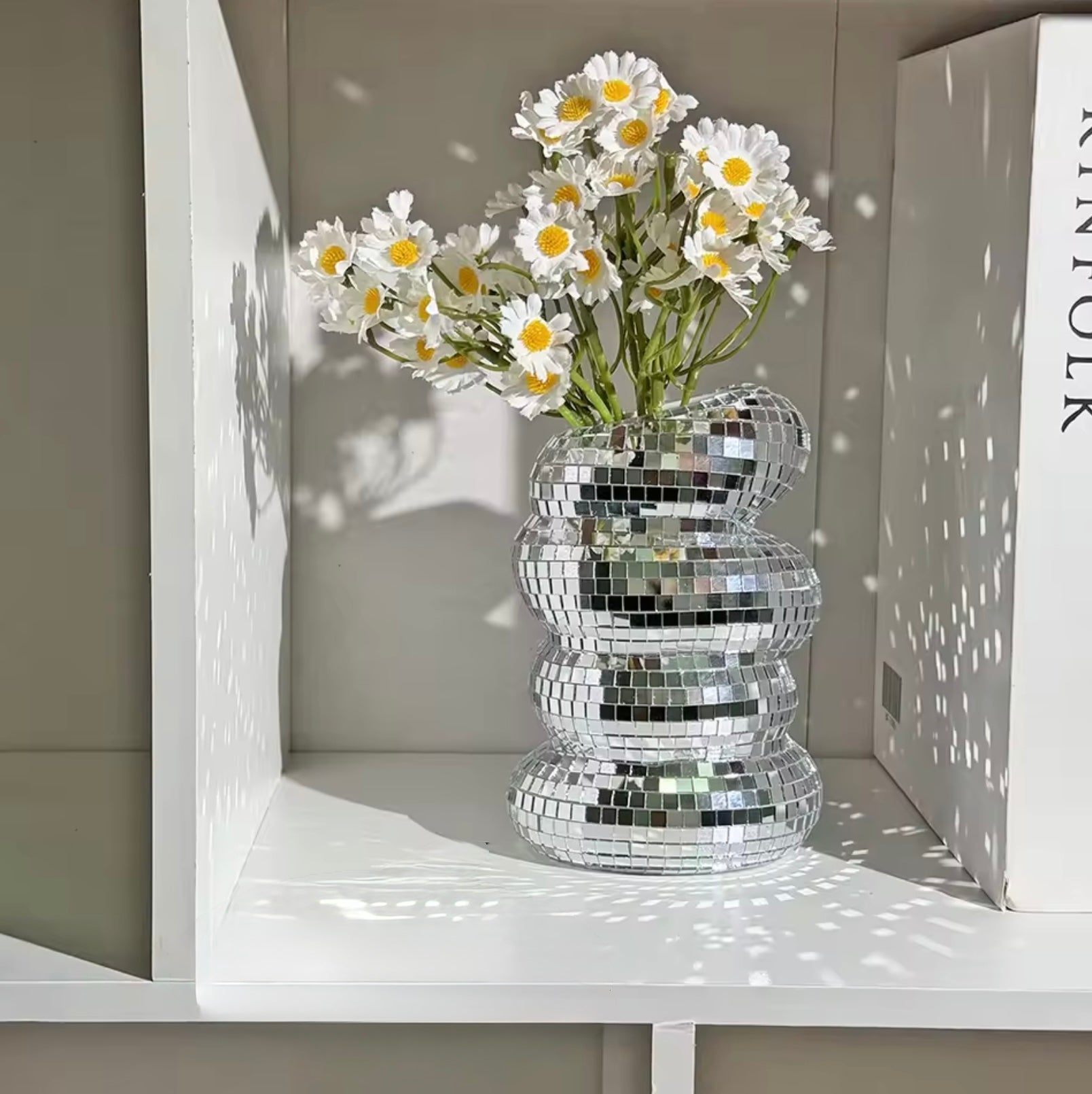 The Plantish Disco Ball Vase placed on a white bookshelf. The vase has daisies inside, and is reflecting the sunlight.