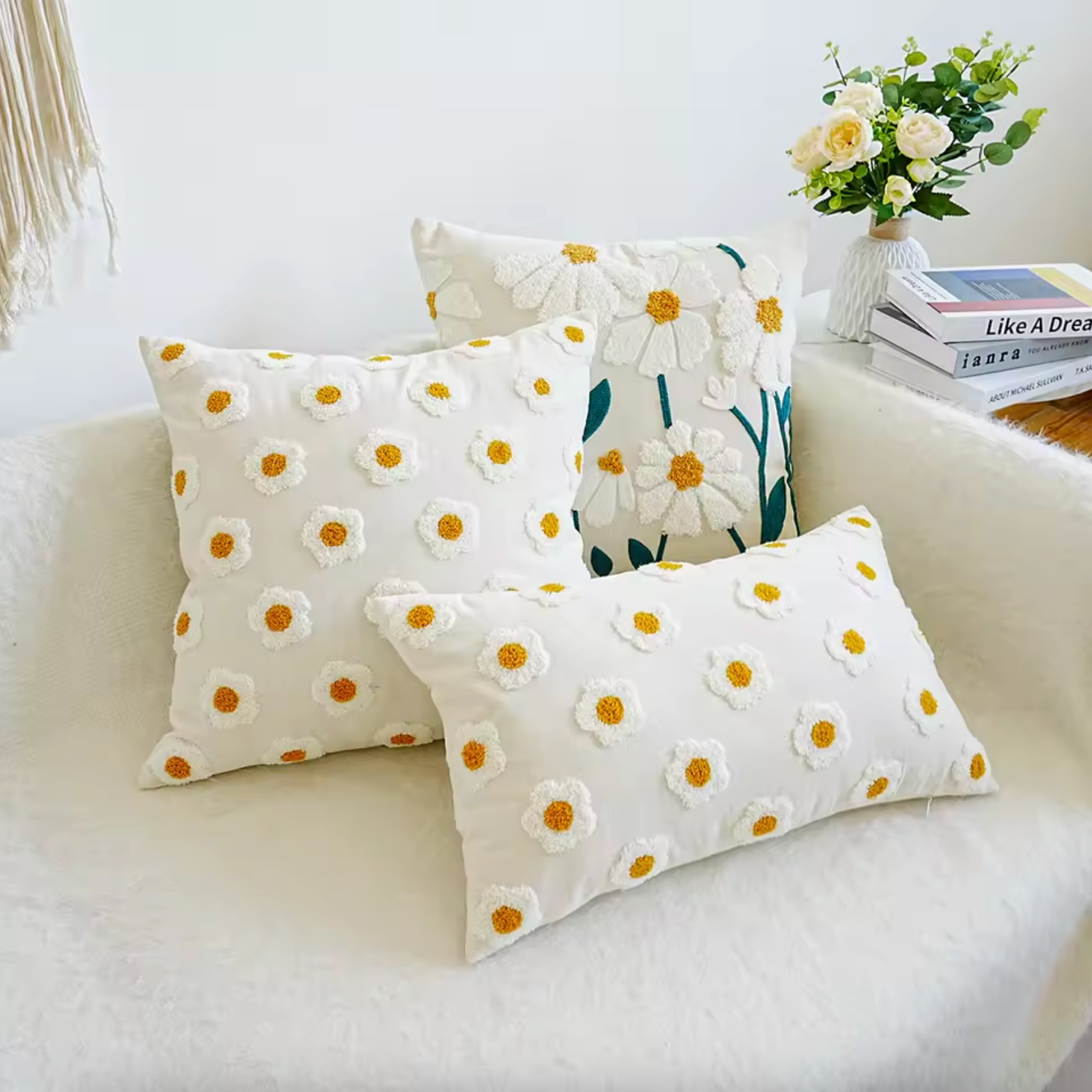 An image showing the Plantish Embroidered Daisy Cushion Cover in three different sizes resting on a white sofa, next to a table with books and flowers on.