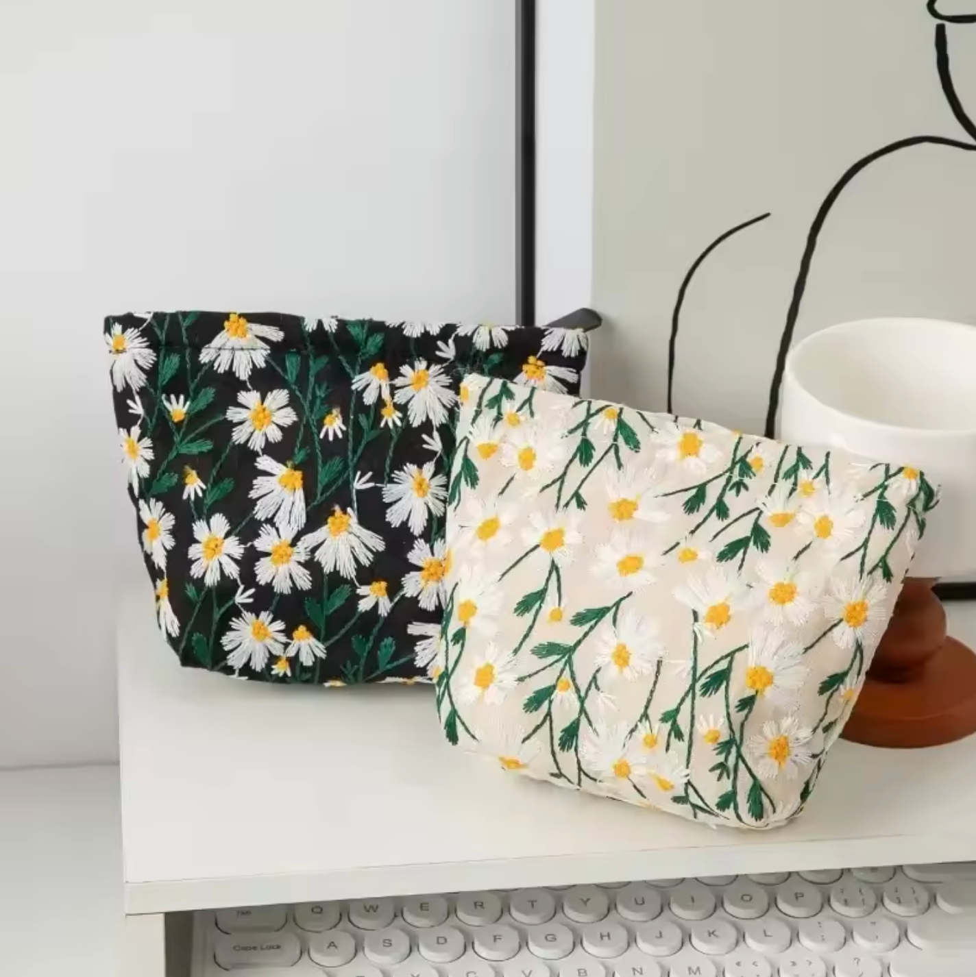 The Plantish Embroidered Little Daisy Makeup Bag 16cm x 6cm x 12.5cm shown in both colours and placed on a white computer desk.