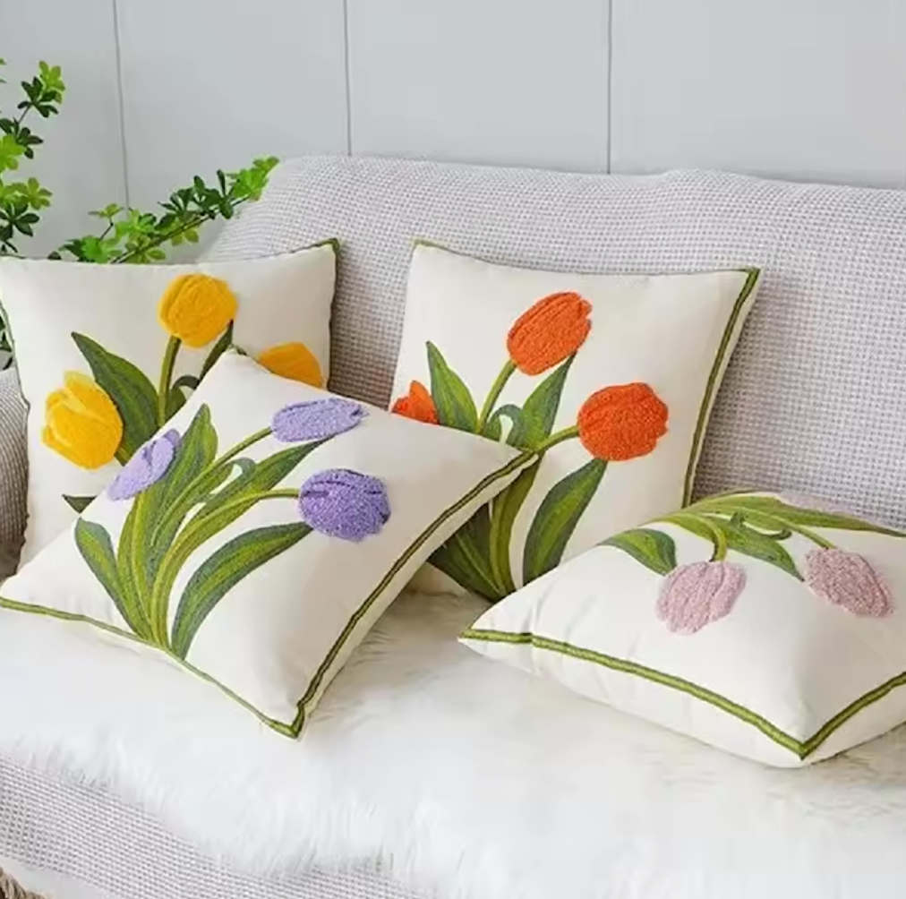 Four designs of the Plantish Embroidered Tulip Cushion Cover, shown positioned on a grey sofa with a white throw, and a green plant visible in the corner. 