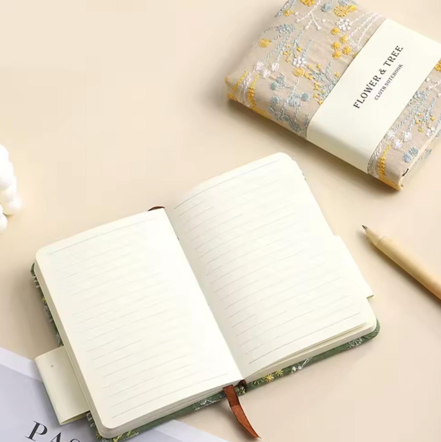 The Embroidered Wildflower Notebook shown in both colours on a cream table. The linen one is closed and the green is open, the lined pages visible.