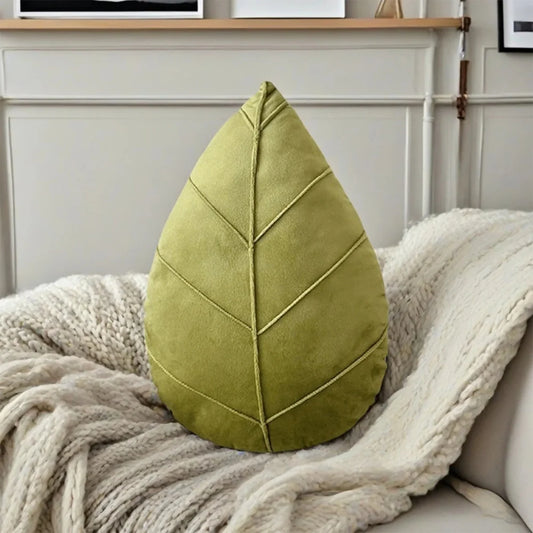 The Plantish Evergreen Plant Leaf Cushion in Light Green postioned on a knitted blanket on a sofa. A living room wall is visible in the background. 