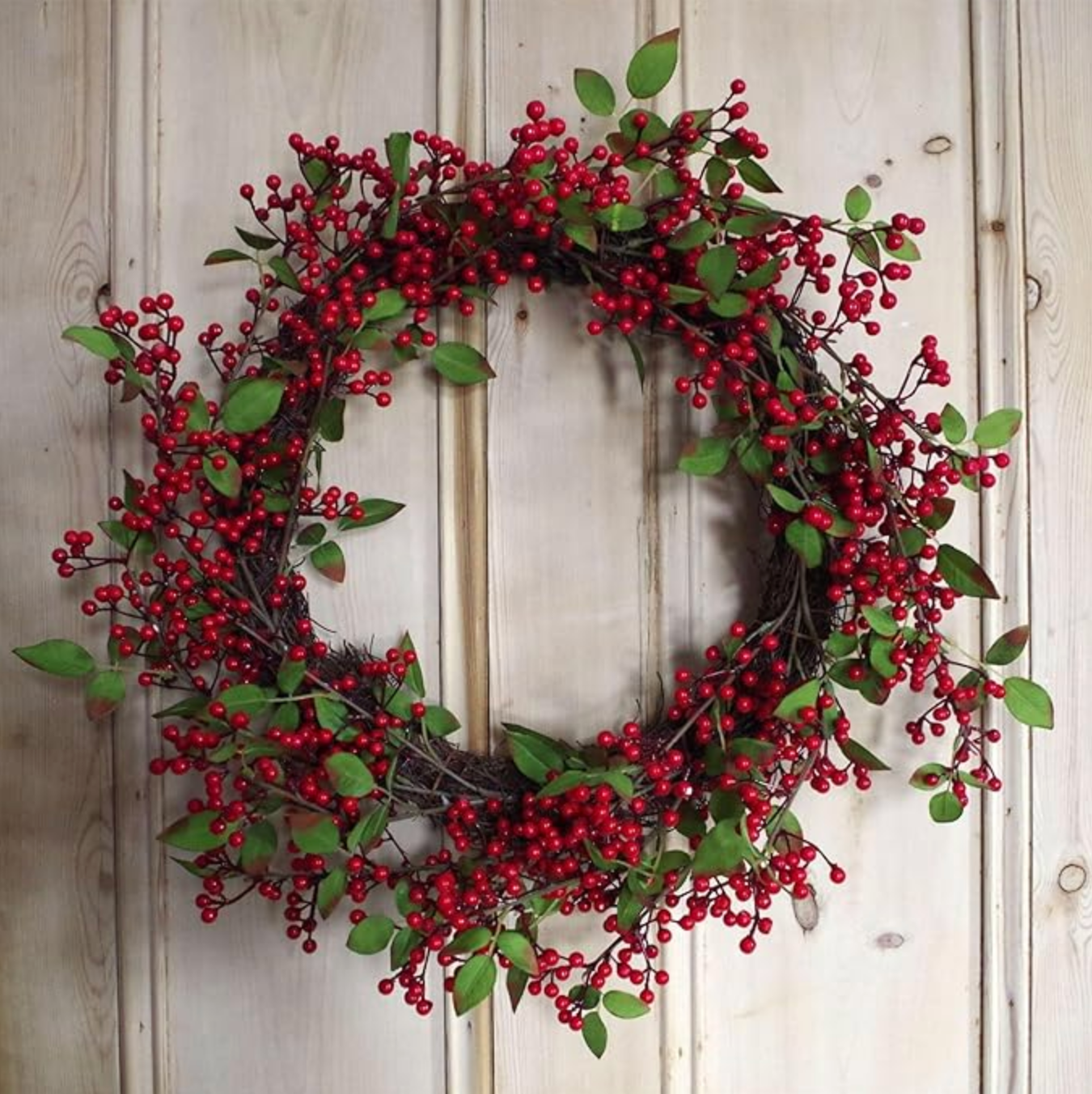 The Plantish Festive Red Berry Wreath hanging on a wooden door, showcasing the red berries, green leaves, and natural woven ring. 