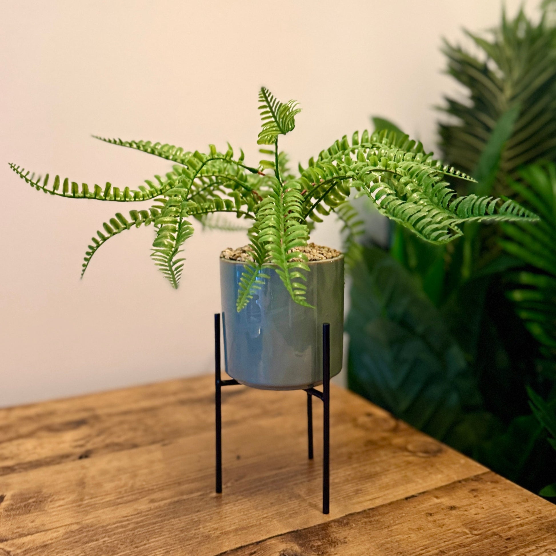 Back view of a Plantish artificial fern in a green ceramic pot with a black stand, showing the base and fixed pebbles, placed on top of a wooden table.