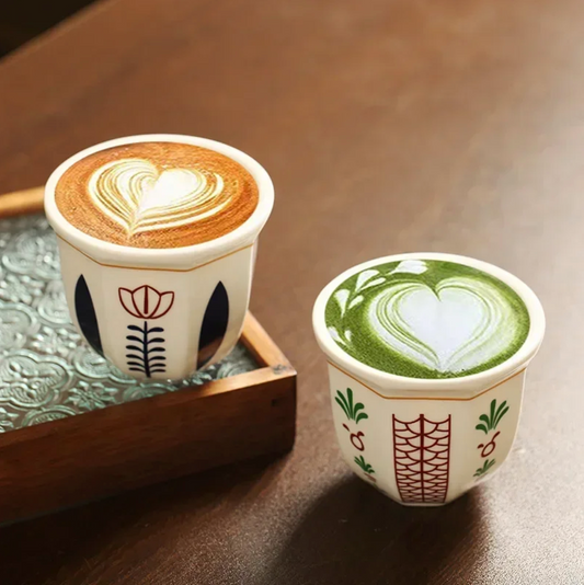 Two Plantish Floral Espresso Cups on a brown table, one with the Leaf design and one with the Flower design. Both have latte art designs and one is on a small tray.