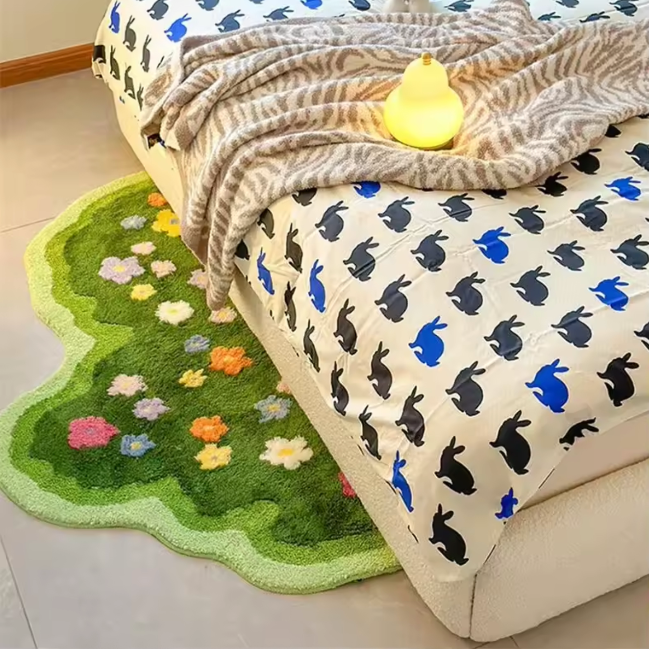 A view of the Plantish Floral Tufted Rug on the floor of a bedroom next to a bed. The bed has a grey and white tiger print blanket with a pear lamp on top.