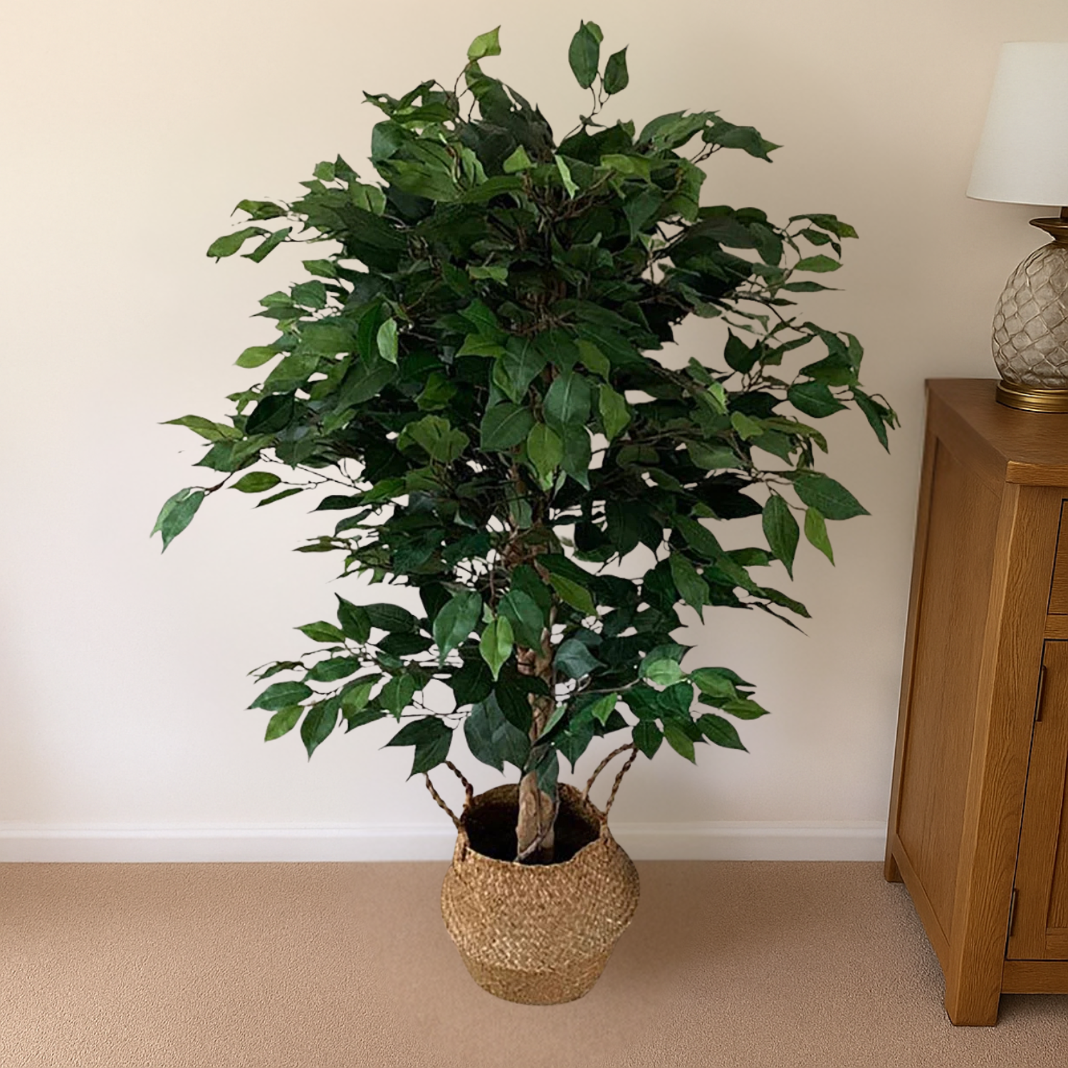The Plantish artificial ficus tree shown placed inside a woven basket that is placed inside a bright living room and a wood cabinet beside it