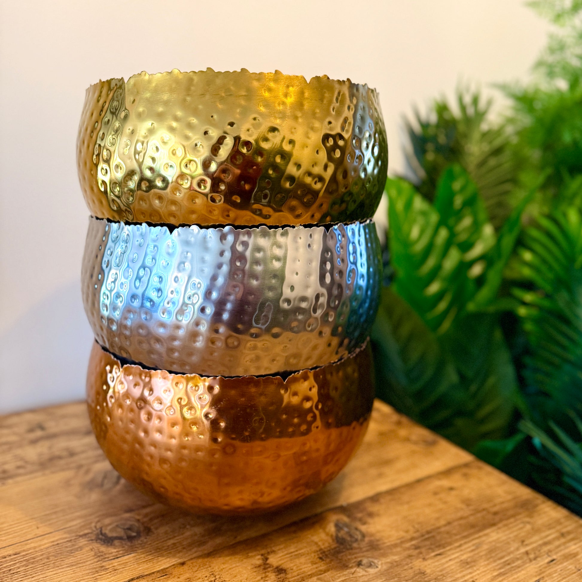 Three metal plant pots in gold, silver, and copper stacked on top of one another on a wood table showing the curved sides and hammered effect.